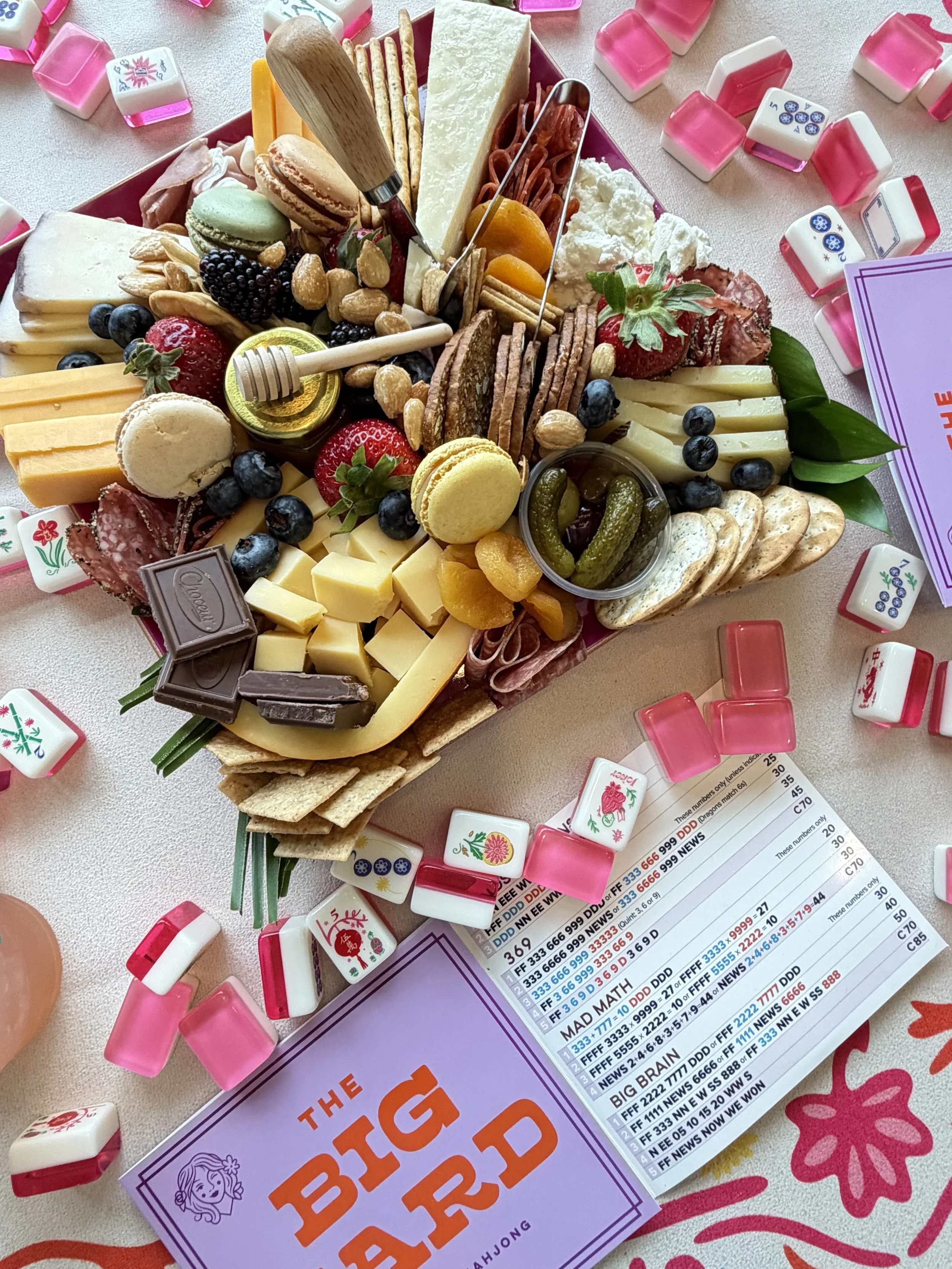 A cheese and charcuterie board with various cheeses, strawberries, blueberries, grapes, nuts, crackers, chocolate, and dried fruit, surrounded by bingo game markers and a bingo scorecard.