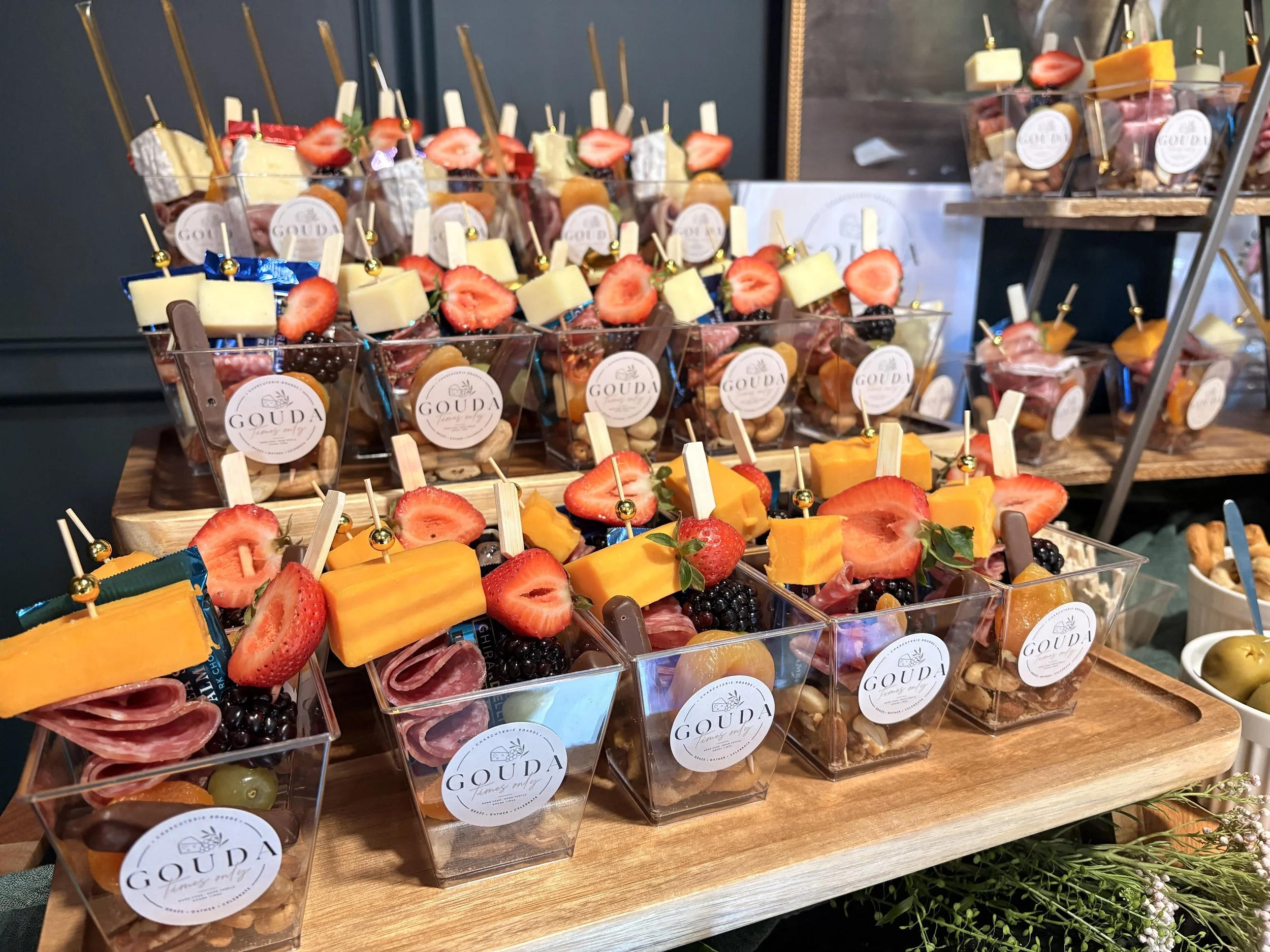 Clear plastic cups filled with an assortment of cheese, strawberries, blackberries, grapes, and slices of orange and other fruits, labeled 'GOUDA' with a logo, arranged on wooden trays and a display table.