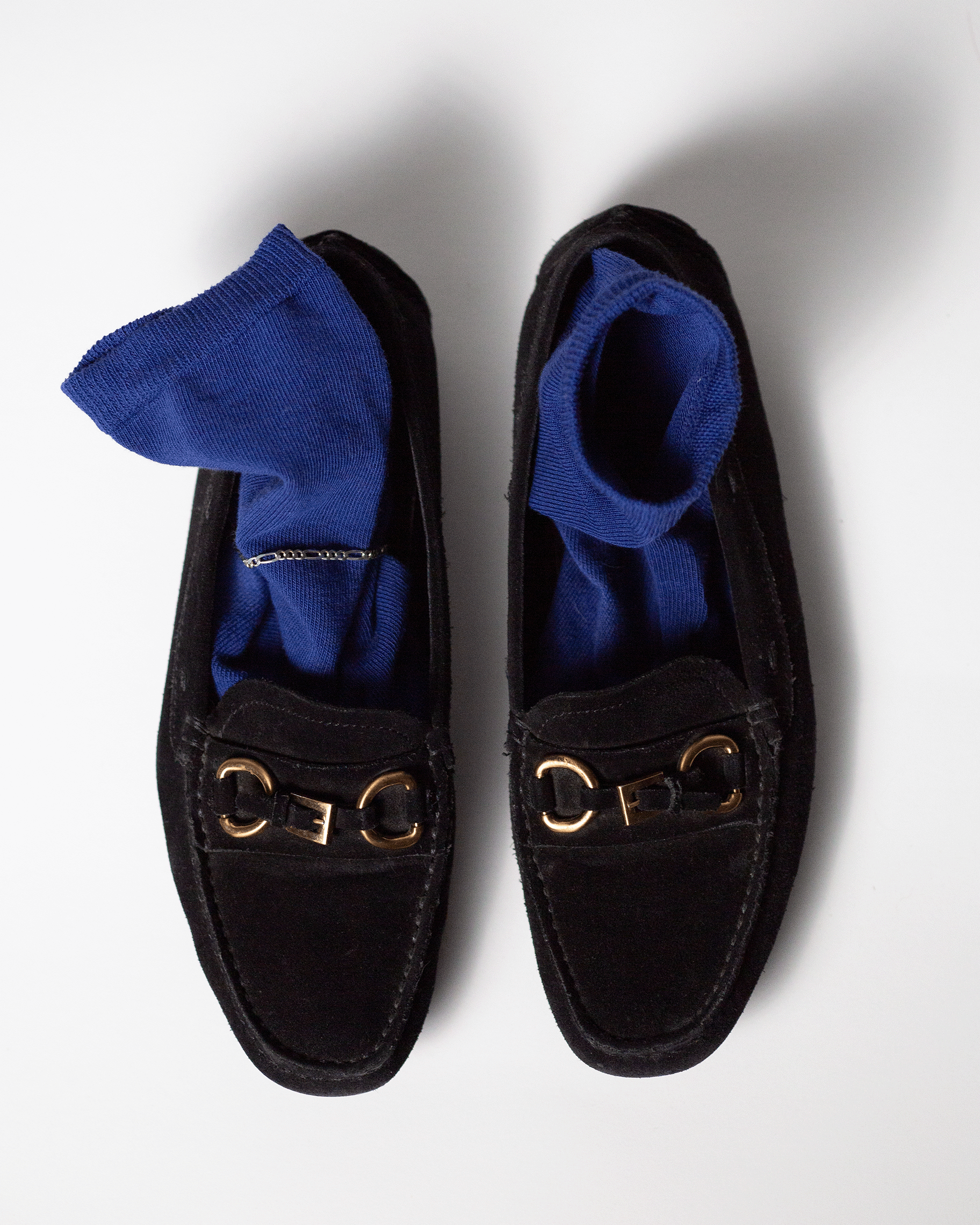 A still life product photograph of pair of black shoes with gold buckle decorations and blue socks, viewed from above on a white surface.
