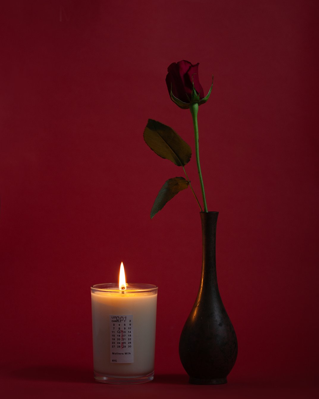 A still life product photograph of a single red rose in a tall, dark vase next to a lit white candle with a margiela inspired label, set against a deep red background.