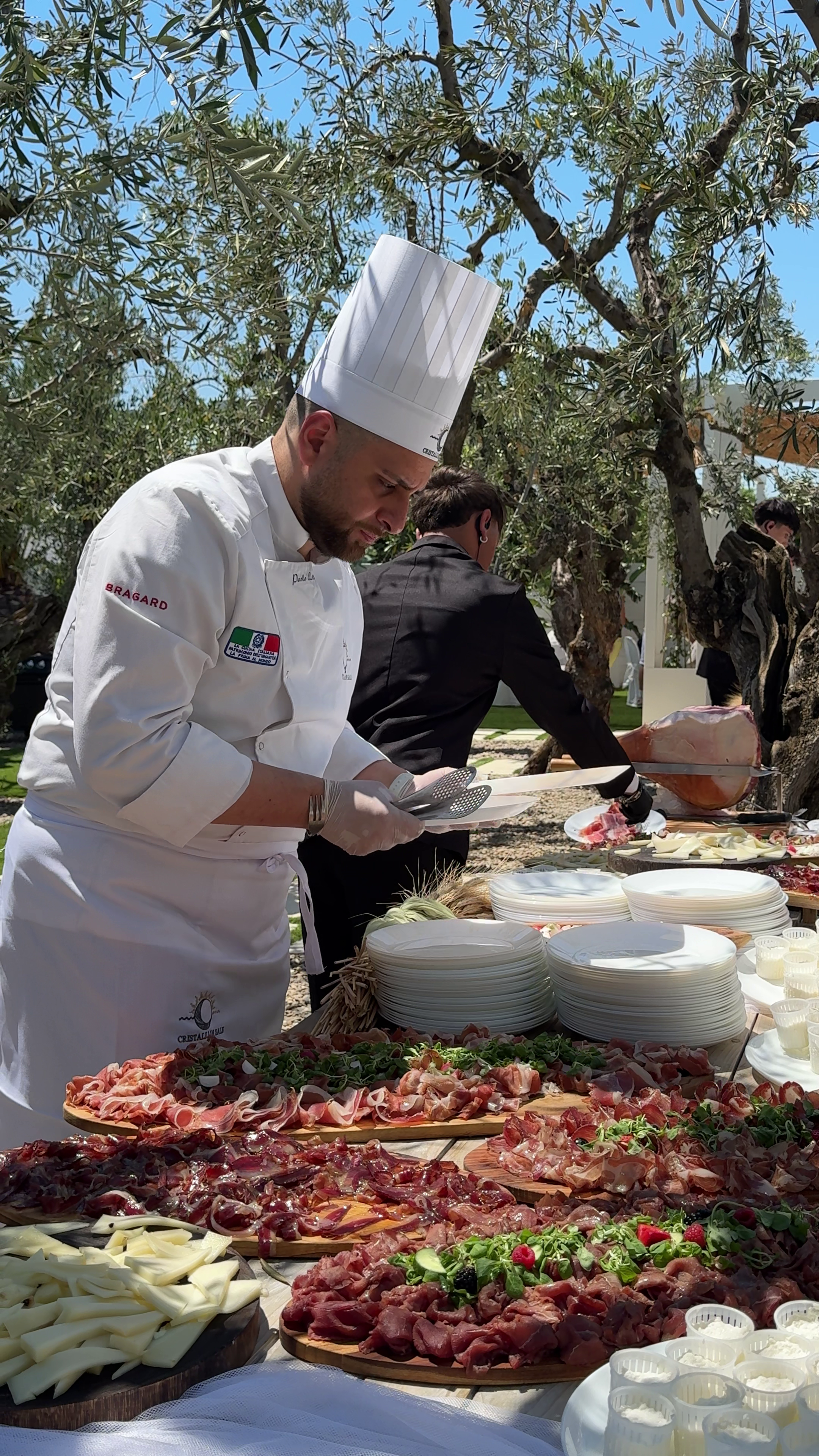 Chef preparing a buffet table outdoors with various meats, cheeses, and garnishes, under a tree with a blue sky.