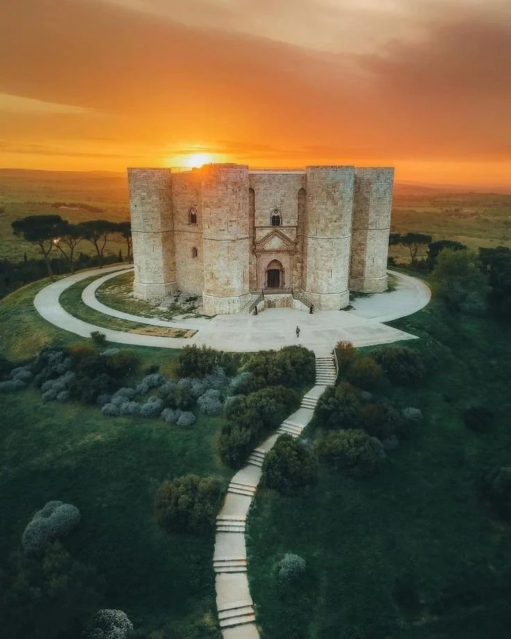 Castel del Monte: A large stone castle on a hill at sunset with a winding pathway leading up to it surrounded by greenery and trees.