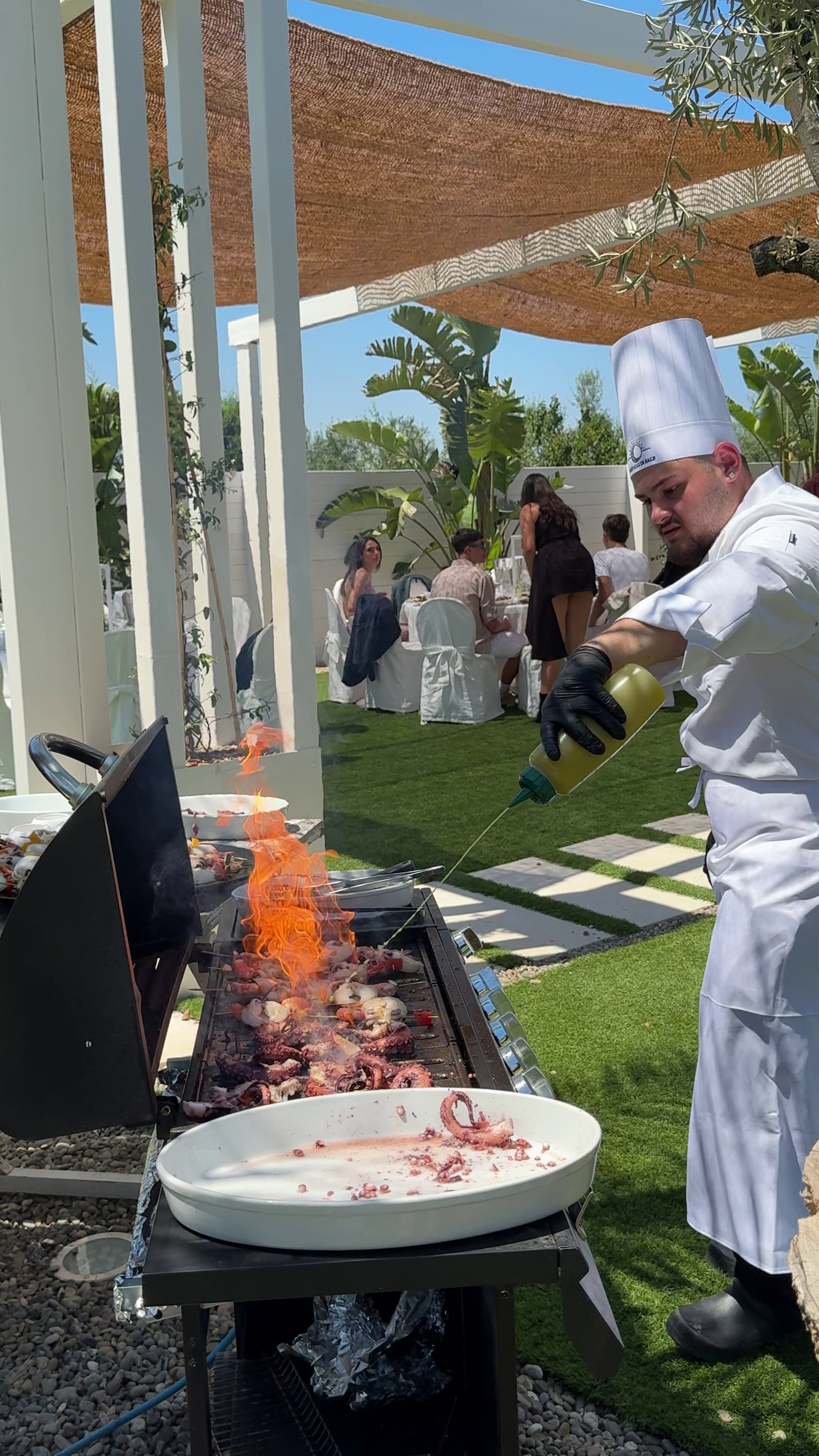Chef grilling seafood, including octopus, on an outdoor barbecue with guests seated at tables in a sunny backyard garden.