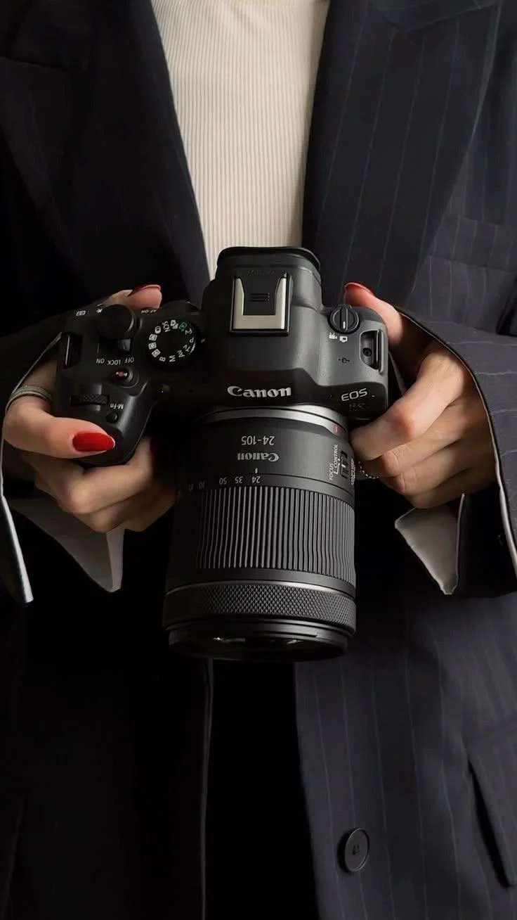 Person holding a Canon EOS camera with a 24-105mm lens, wearing a dark pinstriped suit and a light-colored shirt, with red nail polish.