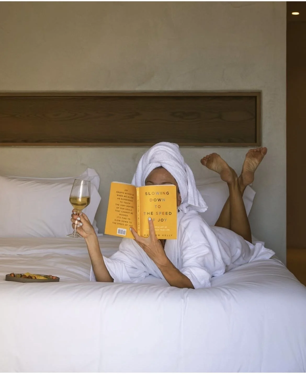 Person relaxing in a hotel bed with a white towel on their head, reading a yellow book titled 'Slowing Down to the Speed of Joy,' holding a glass of white wine, and wearing a white bathrobe.