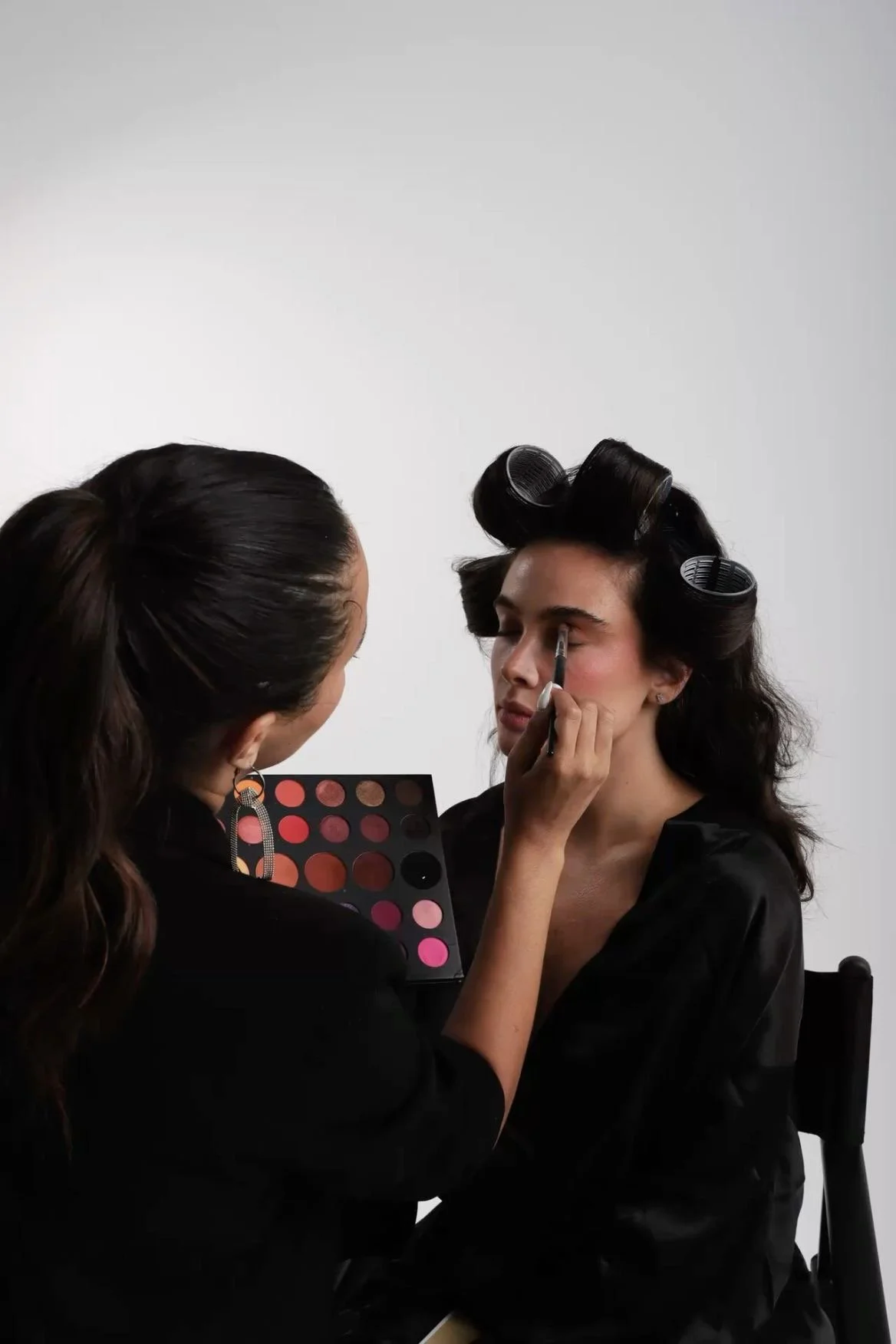 Makeup artist applying eyeshadow to a woman with rollers in her hair, sitting against a plain white background.