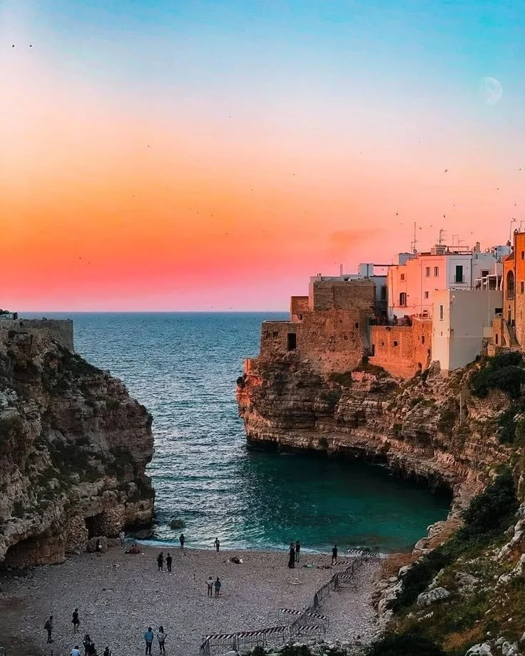 Polignano a Mare: Sunset over a coastal cove with a pebble beach, surrounded by steep cliffs and white buildings, with a small group of people on the beach.