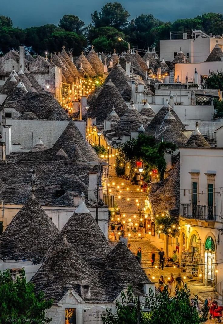 Alberobello: A scenic view of traditional trulli houses with conical roofs decorated with lights, in a picturesque Italian village at dusk.