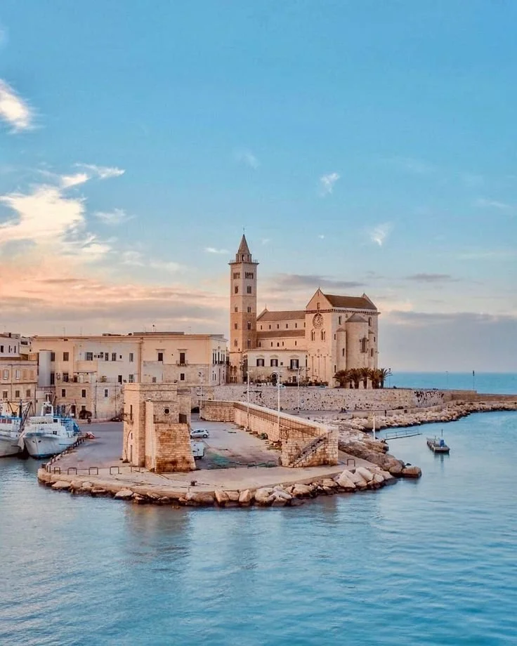 Trani: A coastal scene with a historic stone church and bell tower on a small pier, surrounded by boats and buildings, under a blue sky with some clouds.