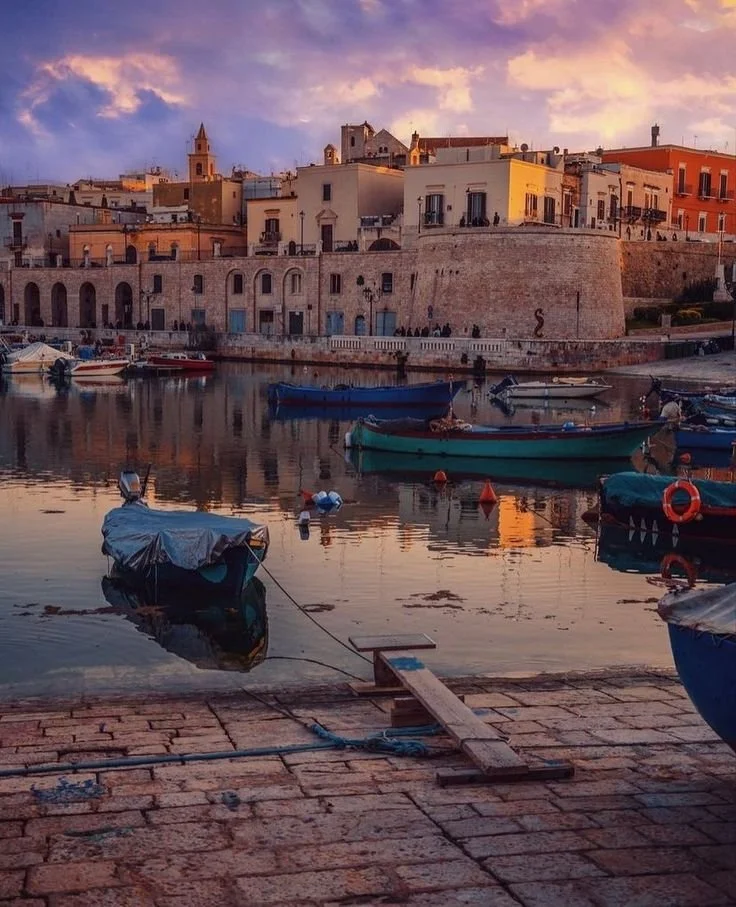 Bisceglie: View of a seaside town at sunset with boats docked in calm water and historic buildings on a hilltop in the background.