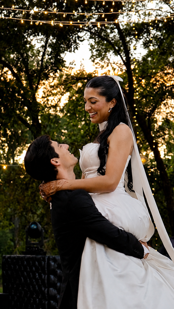 A bride and groom at an outdoor wedding, with the groom lifting the bride in his arms during sunset, surrounded by trees and string lights.