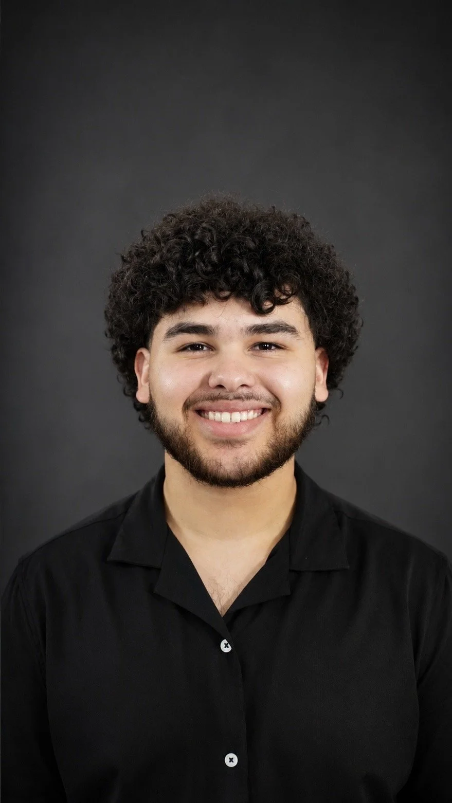 Headshot of a smiling young man with curly hair and a beard, wearing a black shirt against a dark background.