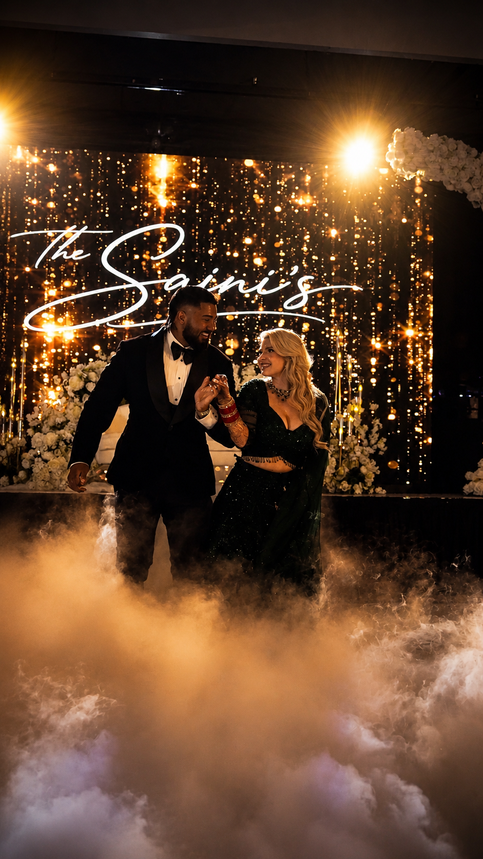 A couple dressed in formal and stylish attire dancing at a wedding reception with a backdrop that says 'The Saine's'. The scene is decorated with white flowers, warm golden lighting, and a fog or smoke effect on the floor.