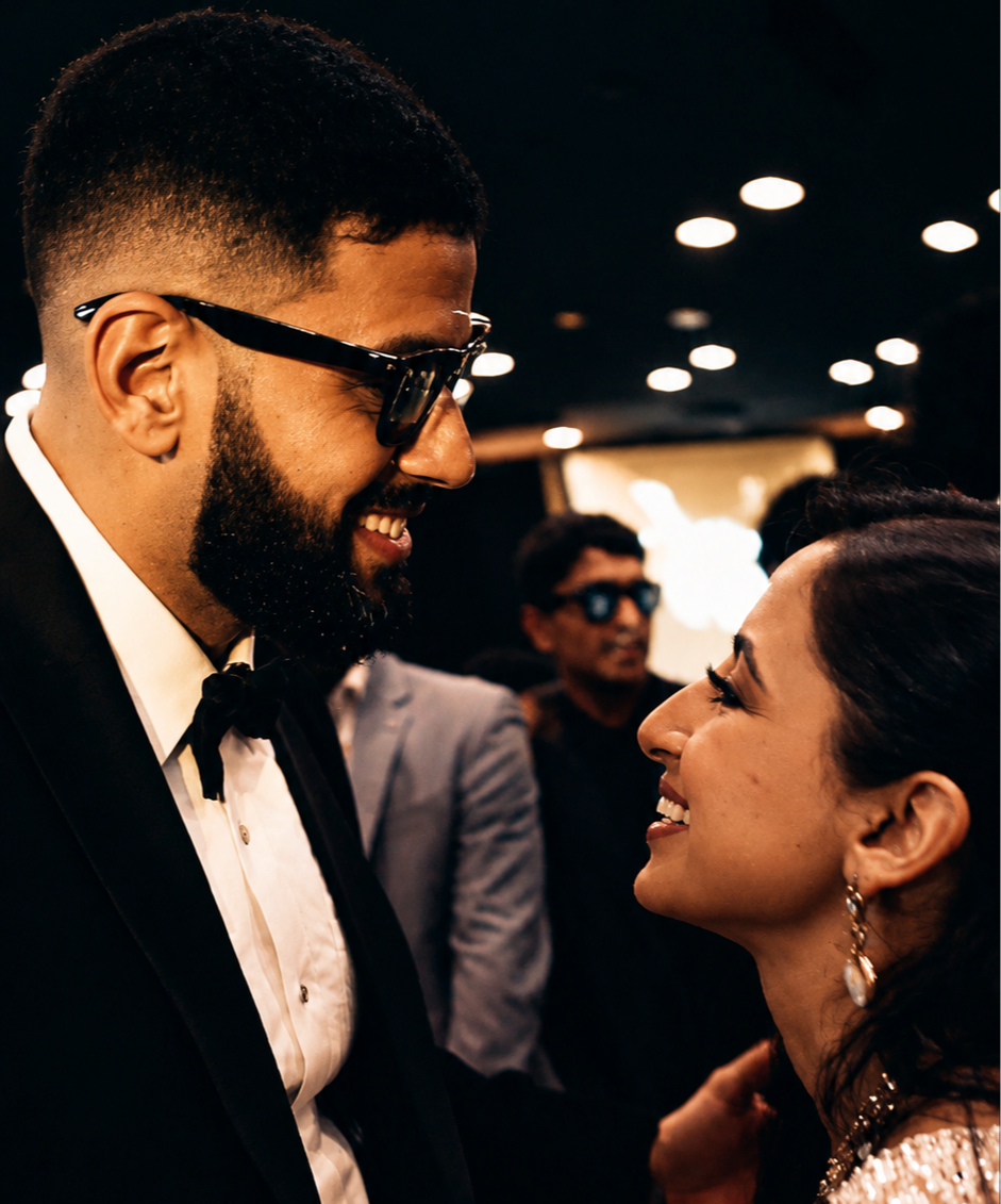 A man and a woman smiling at each other at a formal event, with the man wearing a tuxedo and sunglasses and the woman wearing earrings and a dress, in a dimly lit room with other people in the background.