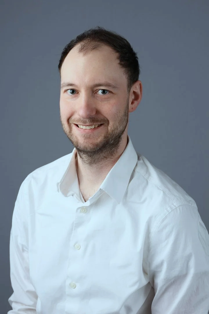 A young man with light skin, dark hair, and a beard, smiling while wearing a white button-up shirt against a plain gray background.