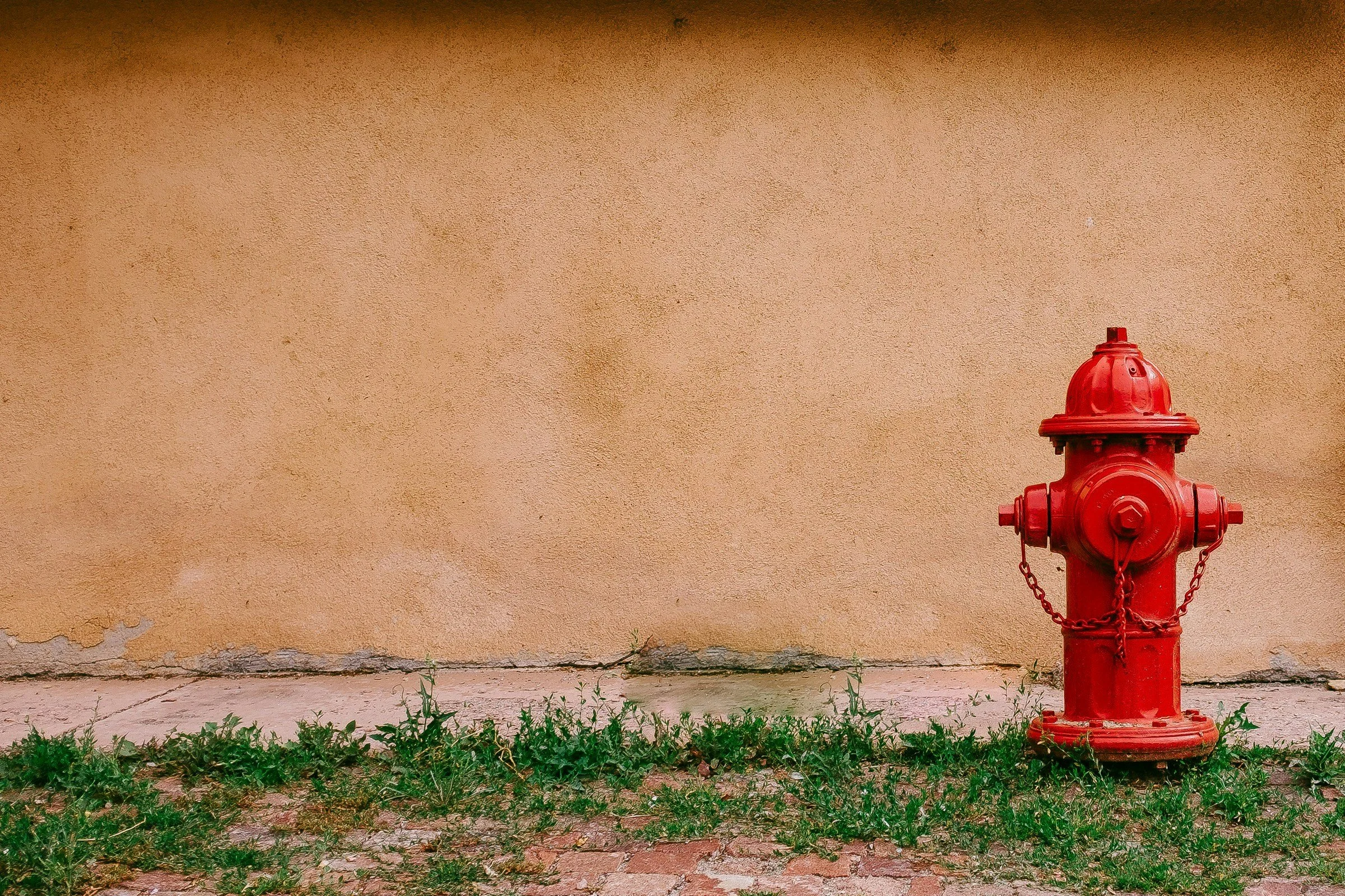 A red fire hydrant with a chain, positioned against a beige wall with peeling paint and a concrete sidewalk with green weeds growing through cracks.