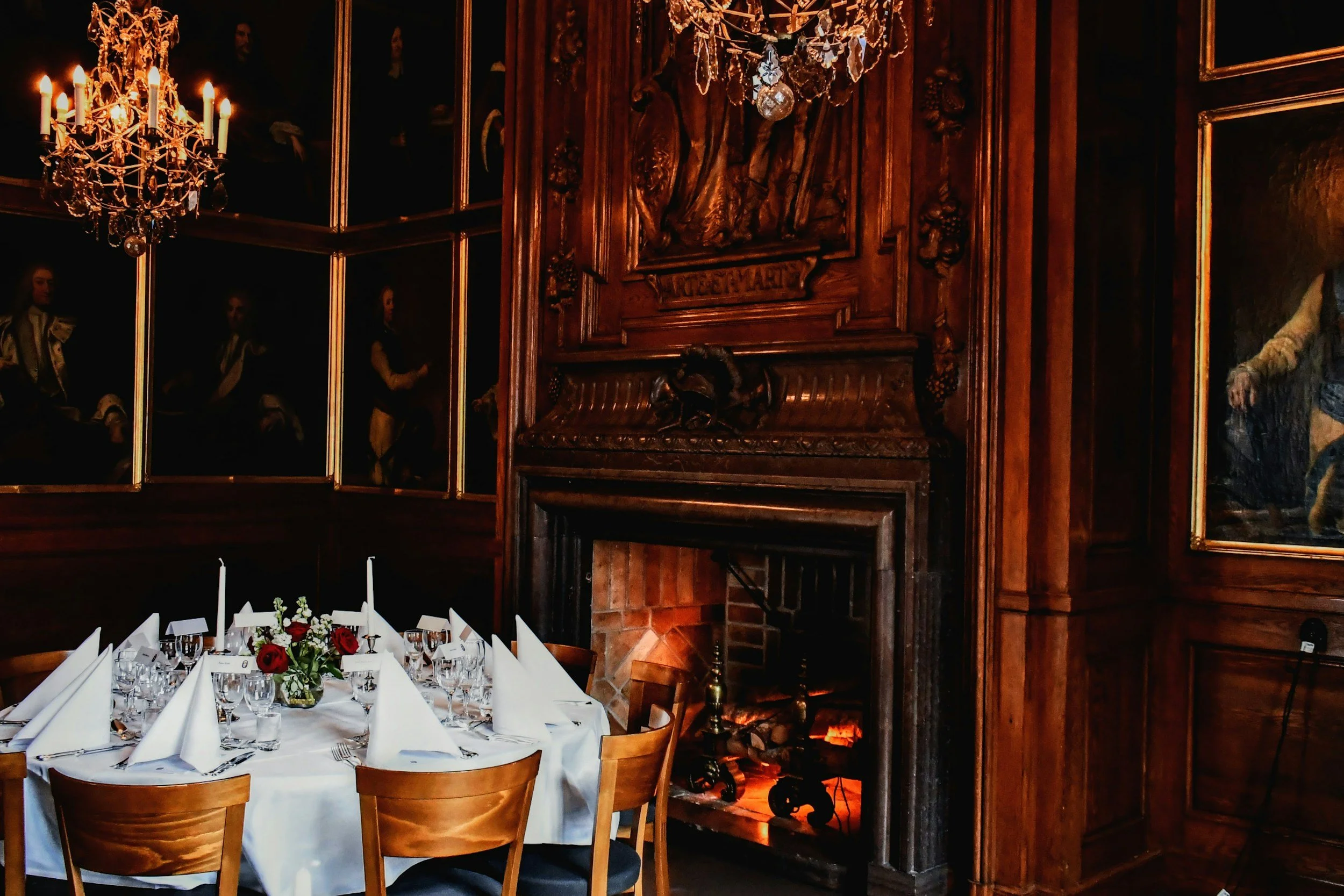 Elegant dining room with a long table set with white napkins, glasses, and floral centerpieces, in front of a fireplace with ornate wooden paneling and chandeliers, with portraits on the walls.