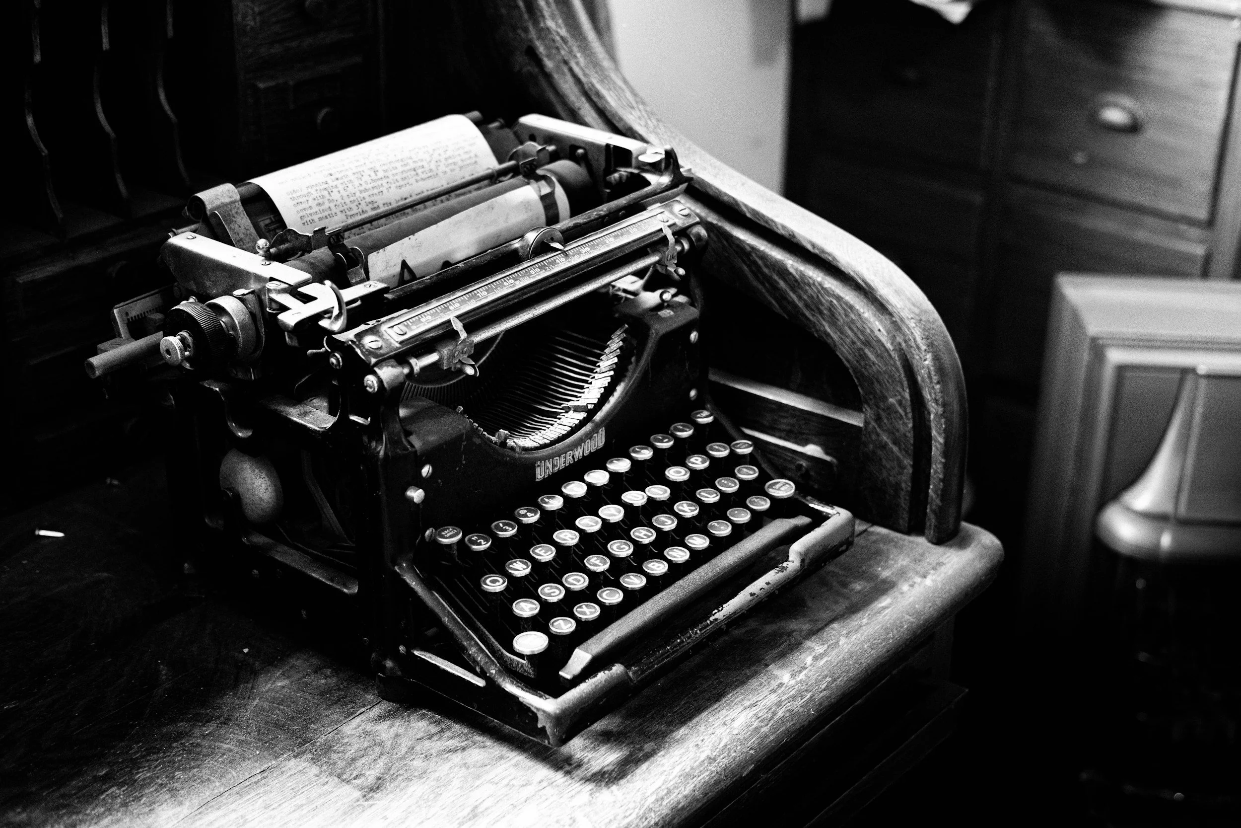 Black and white photo of an old mechanical typewriter on a wooden desk with a drawer in the background.