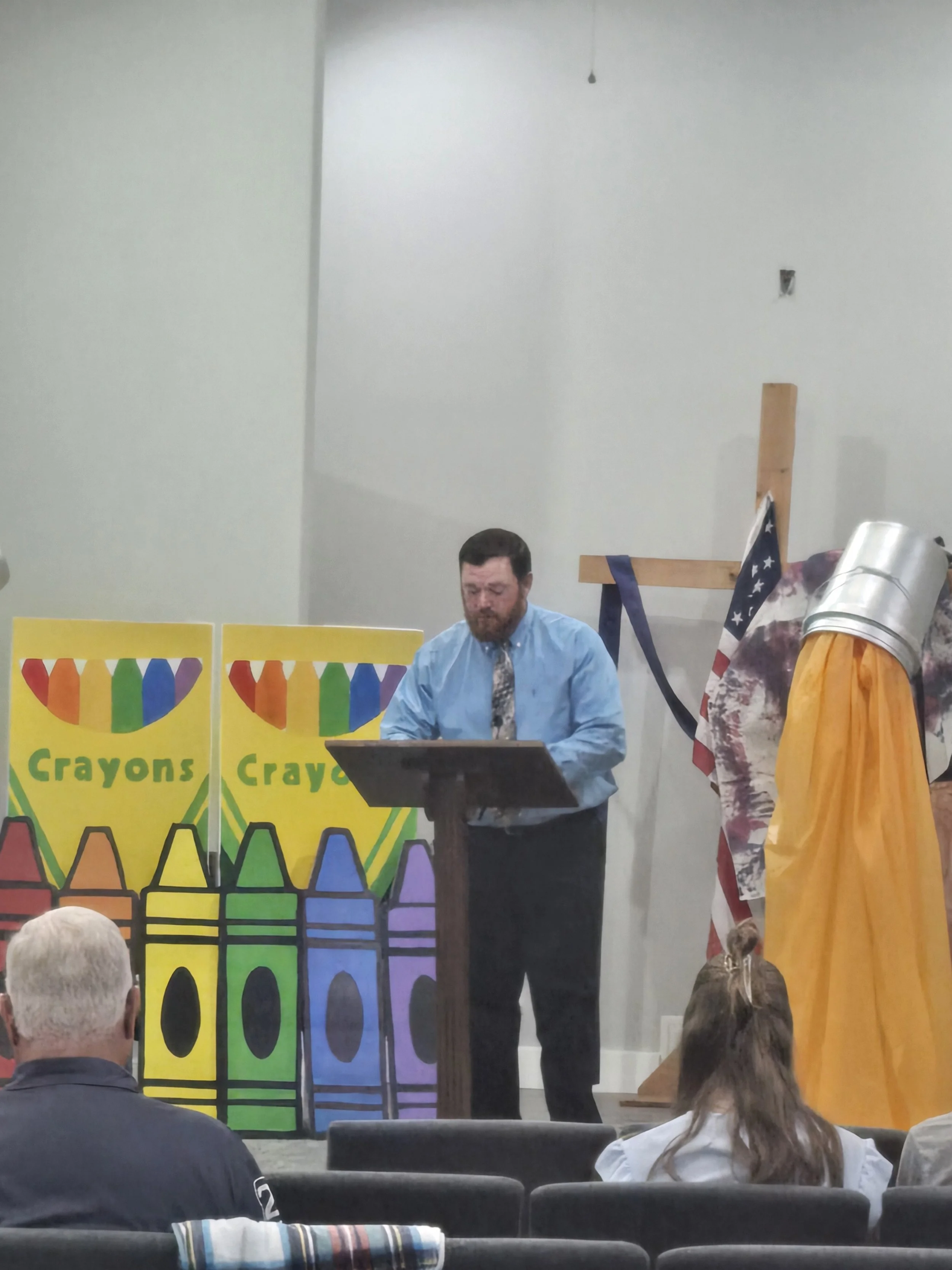 A man in a blue shirt and tie is standing in front of a colorful backdrop featuring large crayons and the words "Crayons" repeatedly. He appears to be speaking or presenting to an audience, which includes children and adults seated and facing the front. There is a wooden cross and an American flag draped over a prop to his right. The setting looks like an indoor event or presentation.