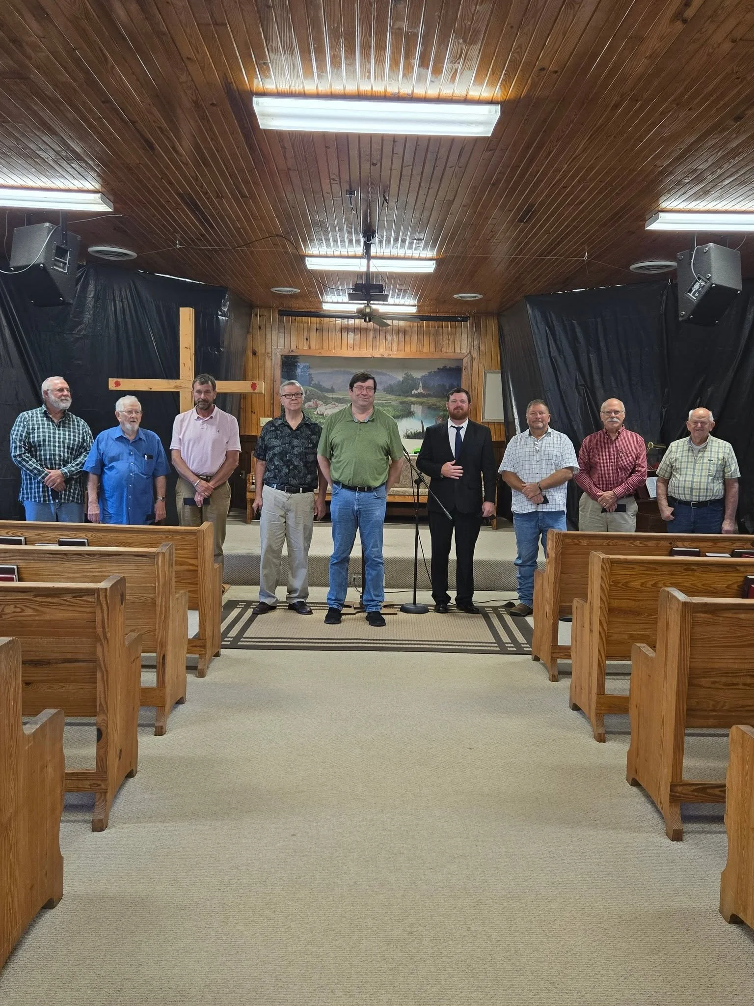 People standing in a church sanctuary with wooden pews, a cross on the wall, and a mural of a lake scene behind the group.