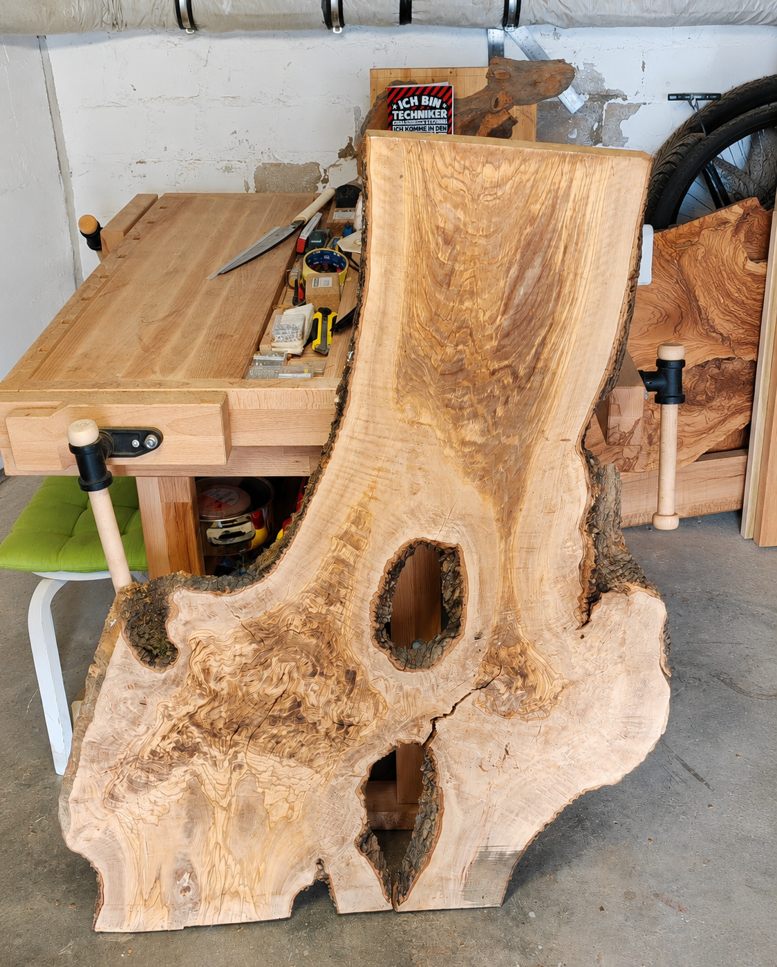 Ein großes Stück Holz mit natürlichen Knoten, das als Tischplatte in einem handwerklichen Arbeitsraum liegt, umgeben von Werkzeugen und Möbeln.