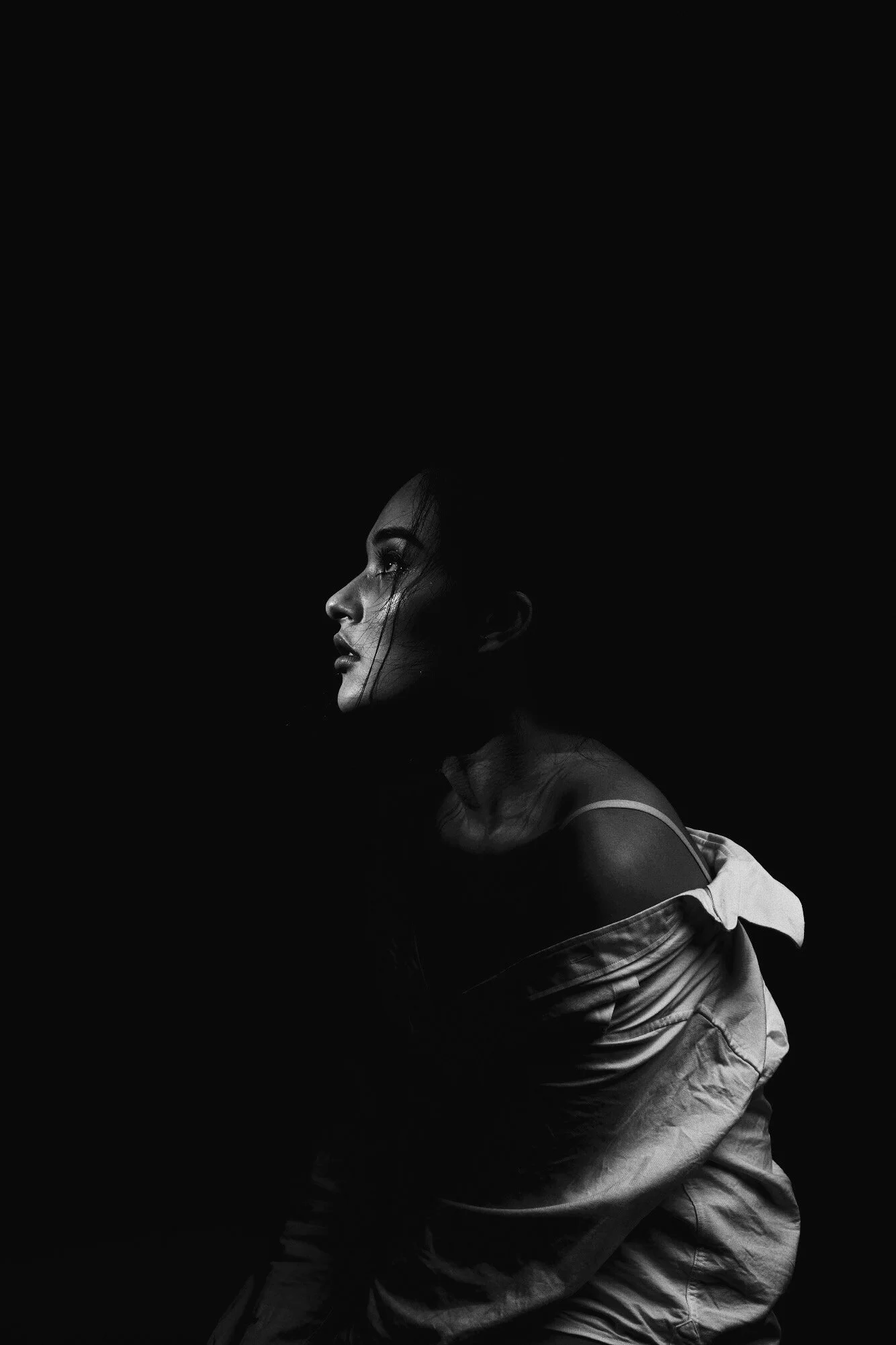 A black-and-white photograph of a woman with dark hair, looking to the left. She is wearing a tank top and loose pants. The background is dark and the focus is on her profile.