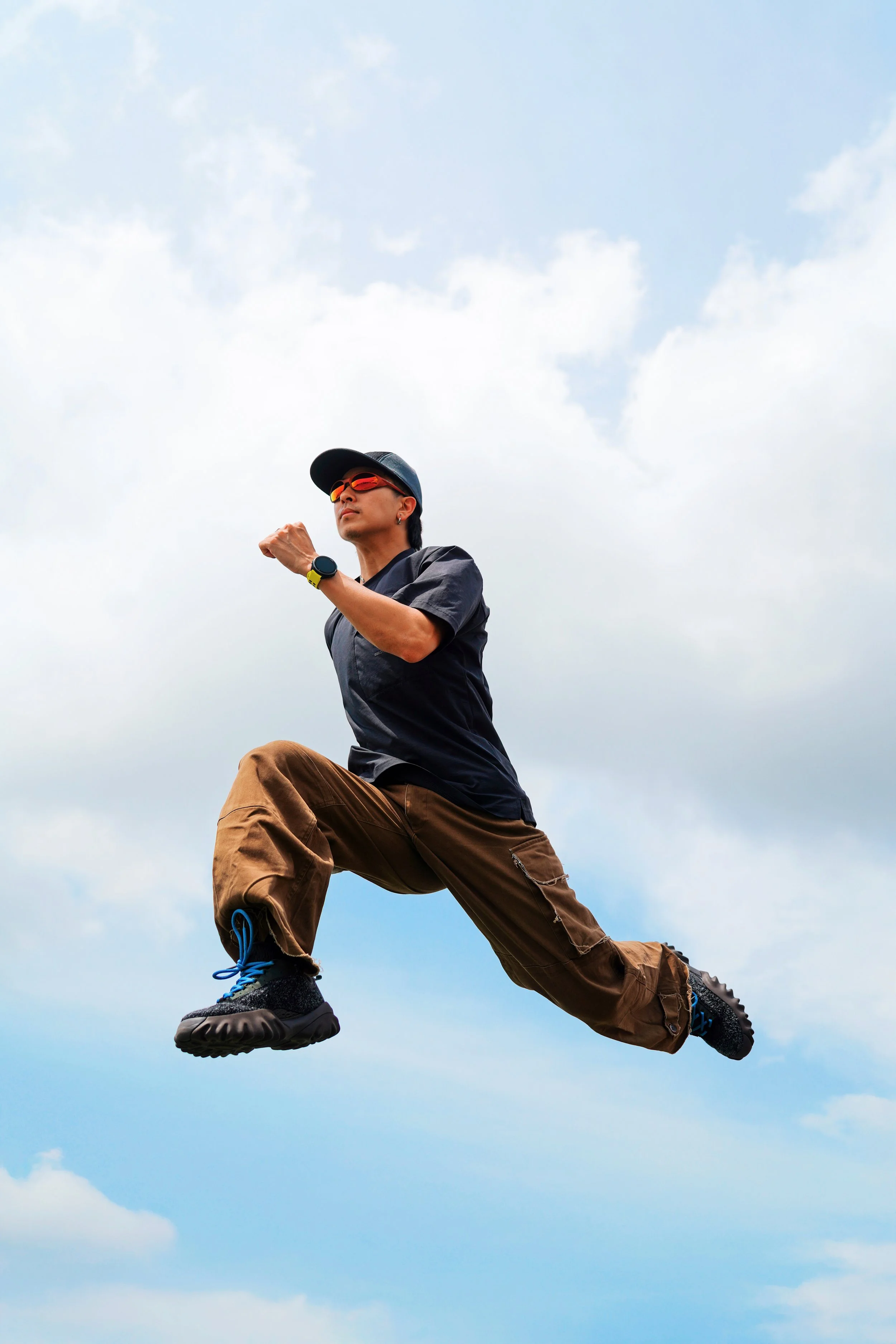 A man dressed in outdoor gear, including a black cap, red sunglasses, black t-shirt, brown cargo pants, and black hiking boots with blue laces, is captured mid-air against a cloudy sky, appearing to leap with one foot in front and the other behind, with a determined expression.