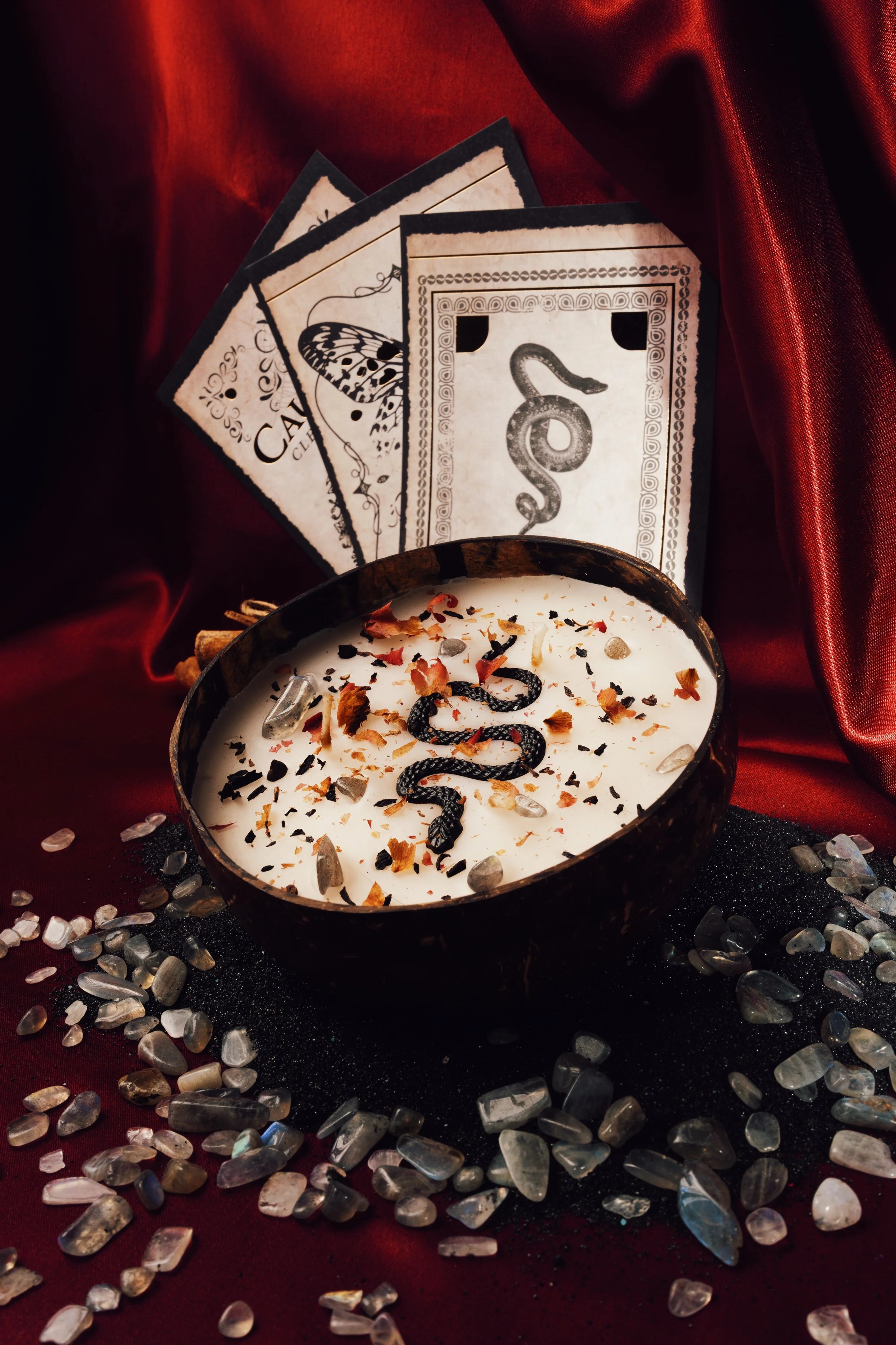 A black bowl filled with a white candle decorated with dried flower petals and a black snake symbol, surrounded by scattered clear and white decorative stones, with a backdrop of red silk fabric and tarot cards with mystical symbols.