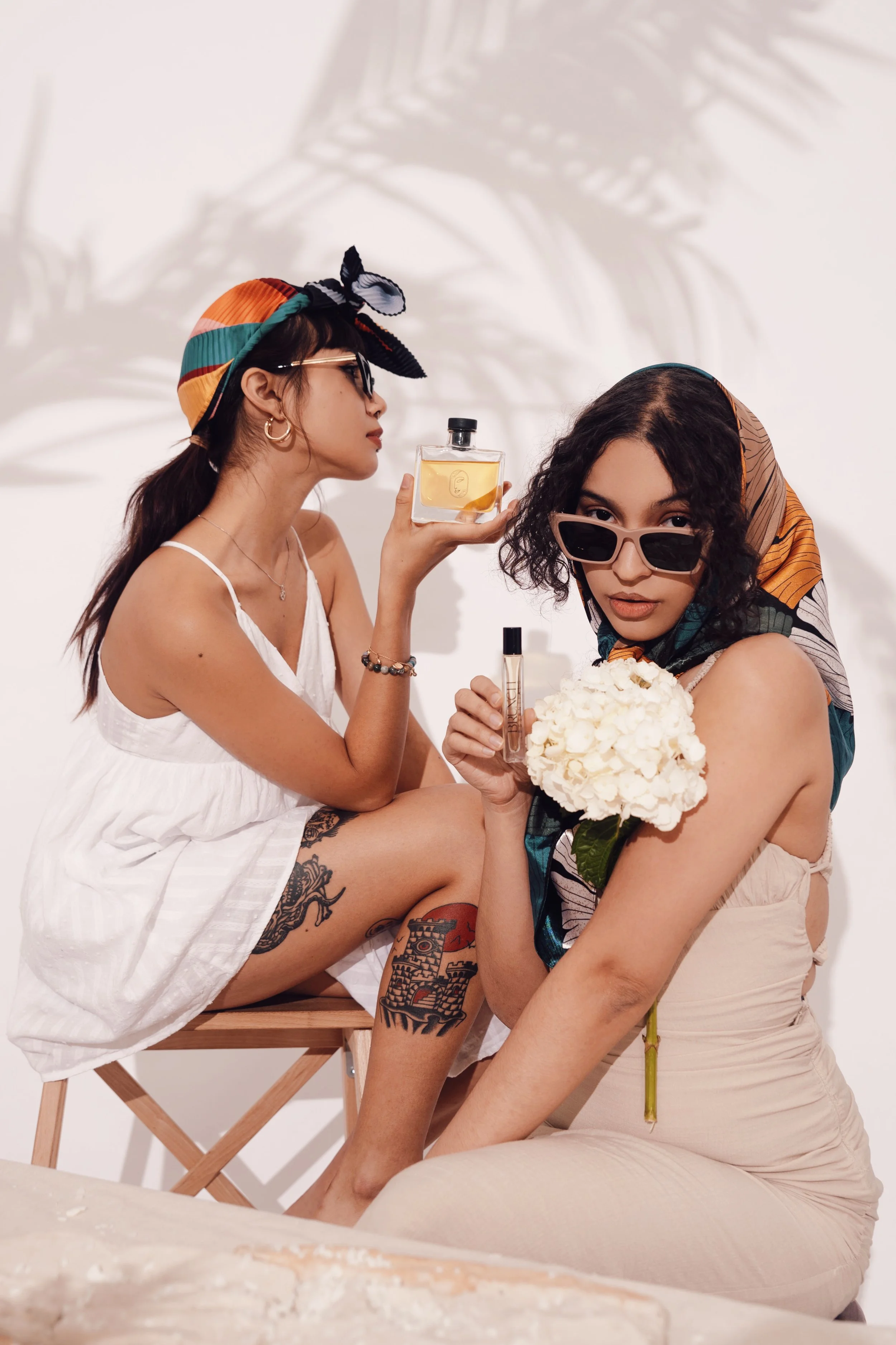 Two women with tattoos and tattoos, wearing sunglasses and headscarves, sitting close together in a minimalistic setting. One woman holds a flower bouquet, and the other holds perfume bottles, with a shadow of a plant cast on the wall behind them.