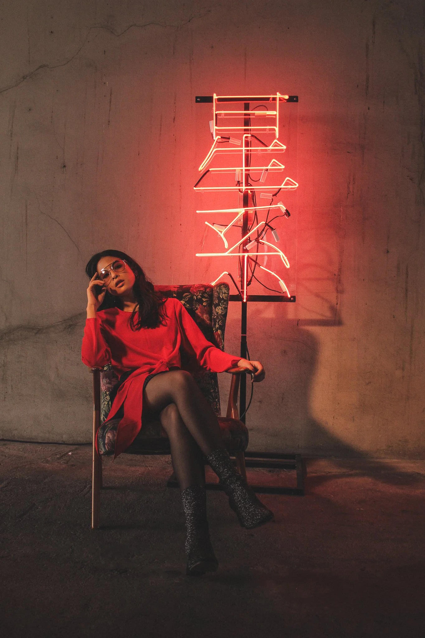 A woman sitting in a vintage armchair against a plain wall with a crack near the top. She is wearing a red dress, glittery high-heeled boots, and glasses, with one hand resting on her head. Behind her is a neon sign with stylized Asian characters, emitting a pinkish-red glow.