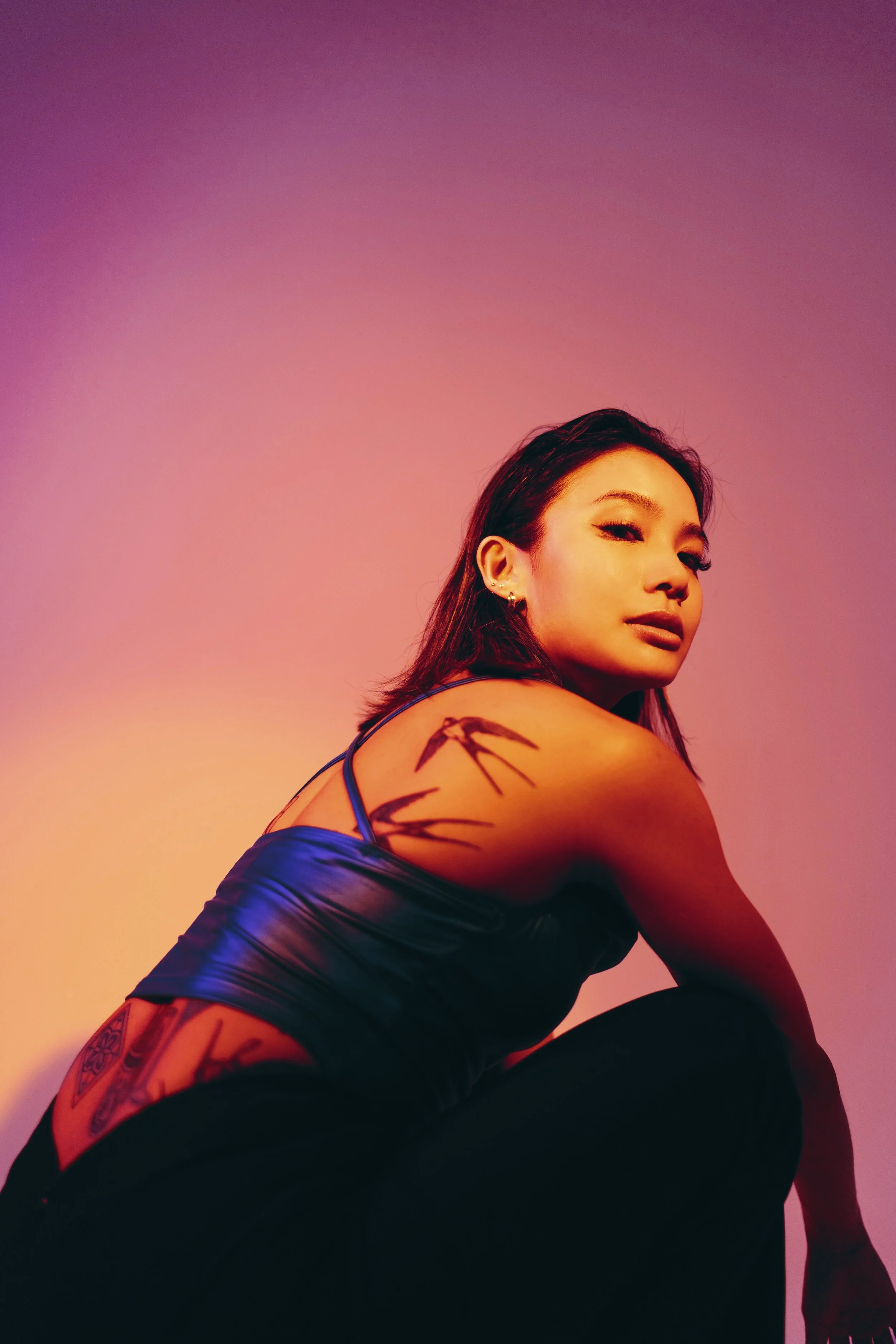 A young woman with dark hair and tattoos on her shoulder and arm, wearing a black top, looking at the camera against a colorful pink, purple, and orange gradient background.