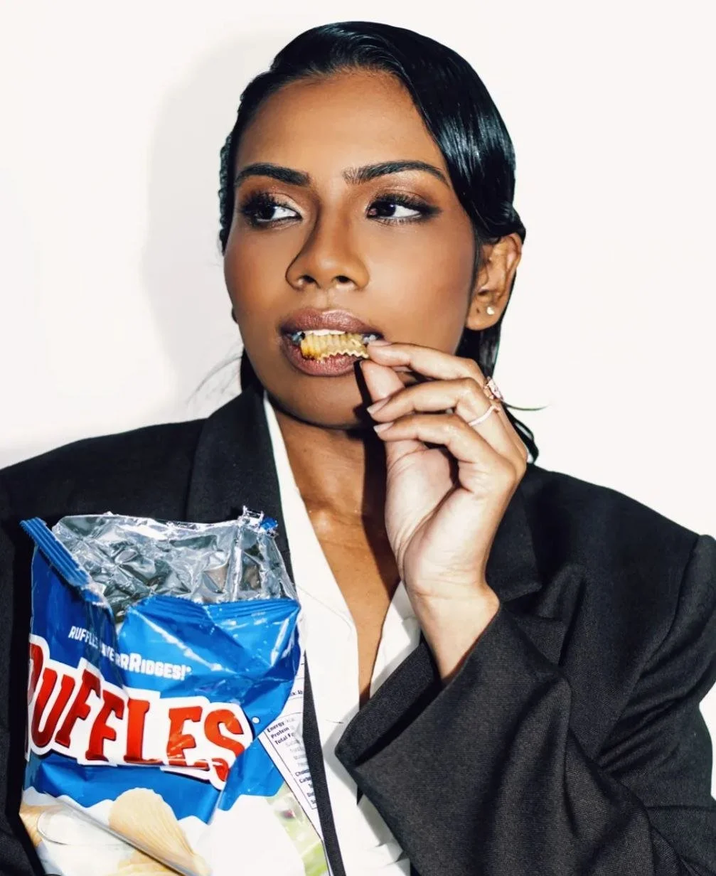 A woman in a black blazer eating a potato chip, holding a bag of potato chips.