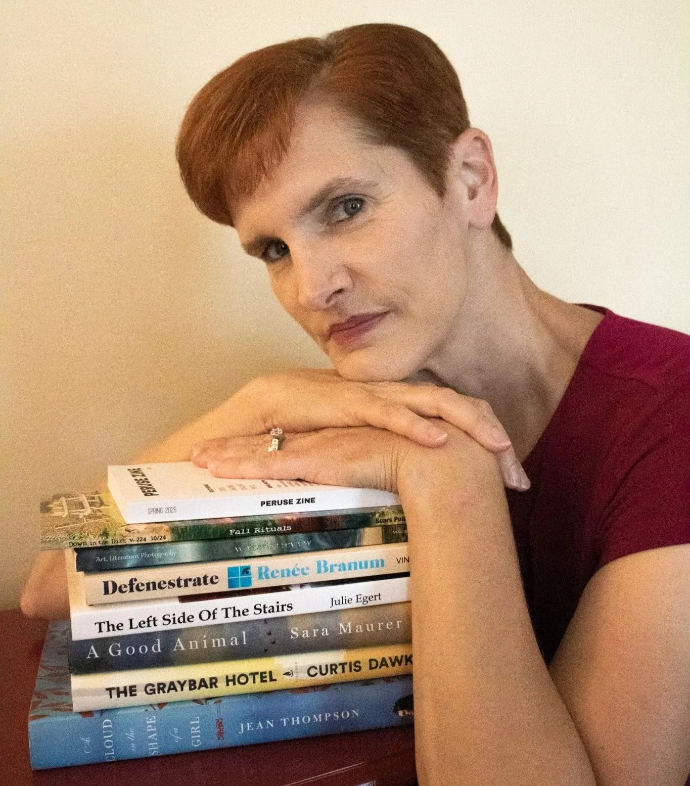 A woman with short red hair and a maroon top is resting her chin on her hands, which are placed on a stack of books and magazines. The books include titles such as 'Defenestrate', 'The Left Side Of The Stairs', and 'A Good Animal'.