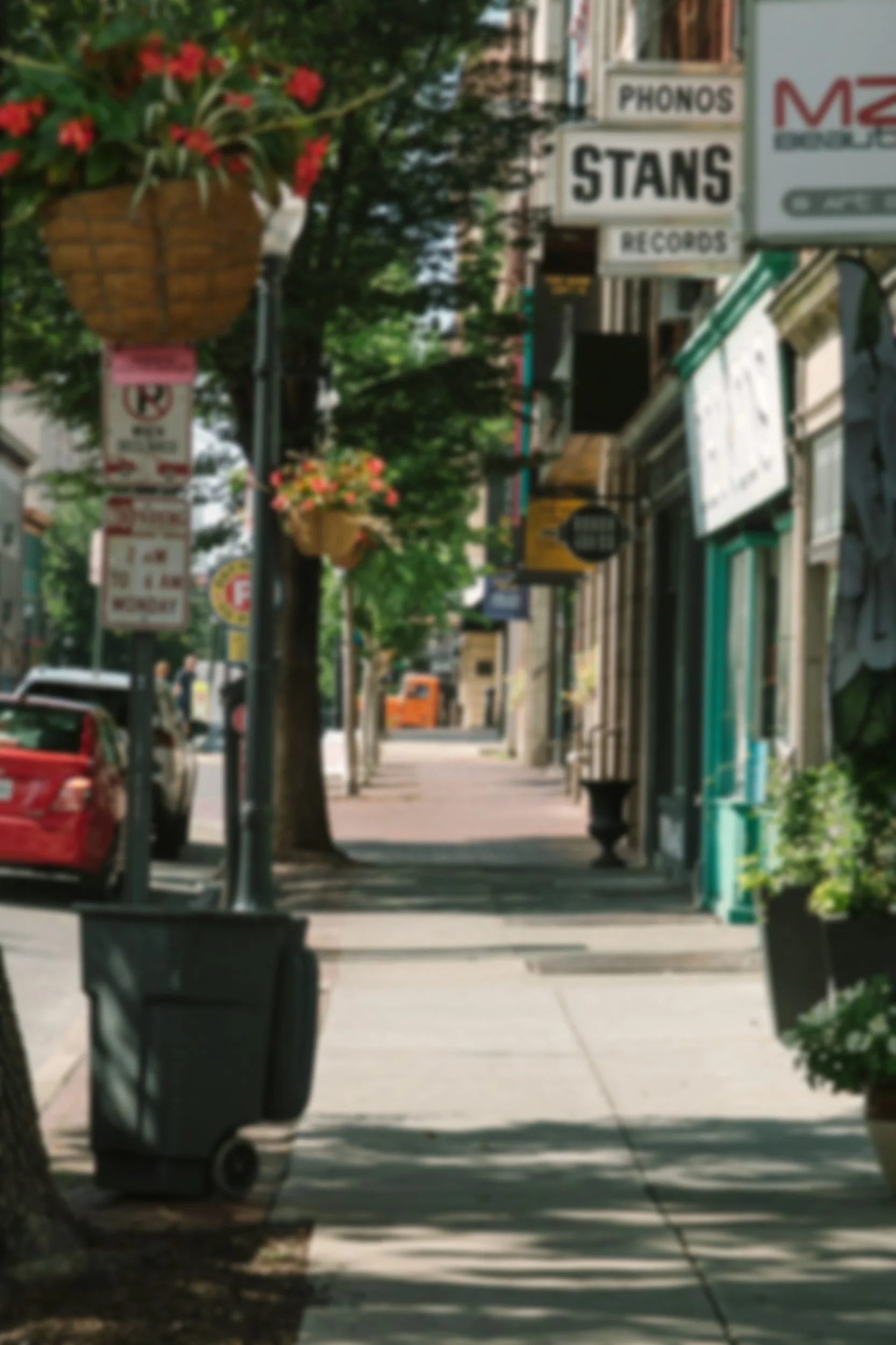 A city sidewalk with hanging flower baskets, several signs, and a trash bin in the foreground. Cars are parked along the street, and trees line the sidewalk. The buildings have storefront signs.