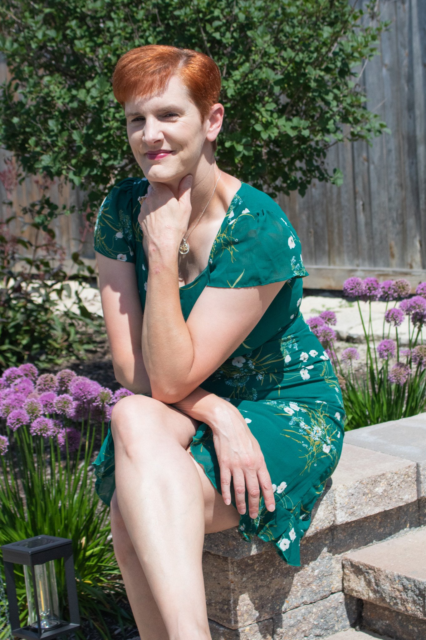 A woman with short red hair sitting outdoors on a stone bench, wearing a green floral dress, with her hand on her neck and her left knee exposed. She is surrounded by purple flowers and greenery and is smiling softly at the camera.
