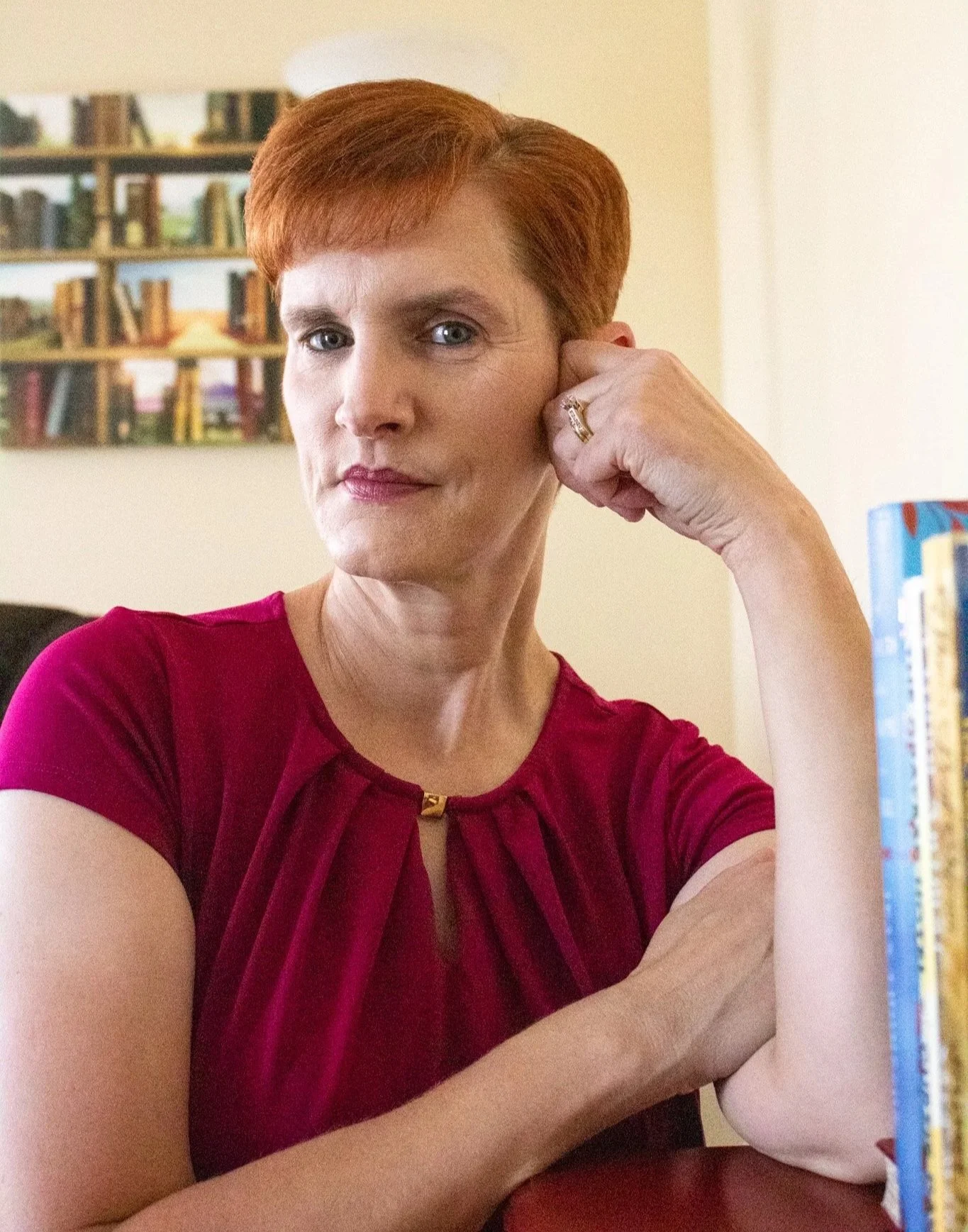 Woman with short red hair wearing a red dress, sitting at a table, with bookshelves in the background.