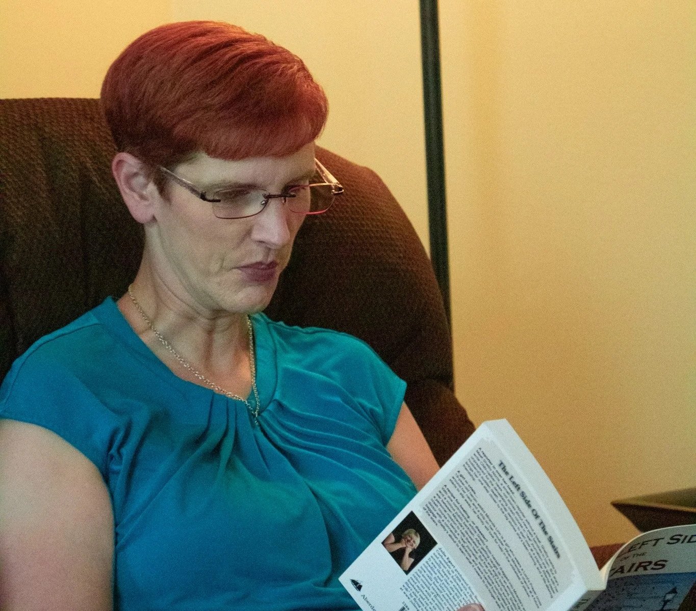 A woman with short red hair, wearing glasses and a blue top, sitting in a brown armchair, reading a pamphlet.