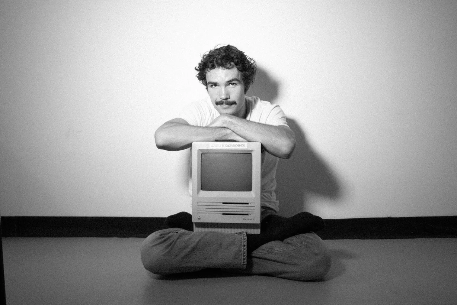 Black and white photo of a man with curly hair and a mustache, sitting cross-legged on the floor, resting his arms on an old computer monitor, with a plain wall background.