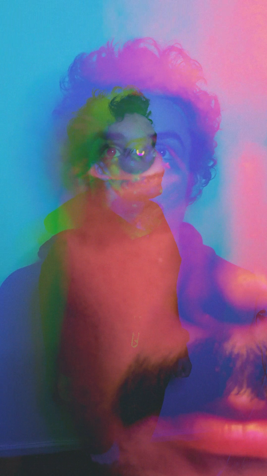 A colorful, layered image of a person's face and a larger face in the background, with vibrant hues of pink, blue, green, and purple creating an abstract, surreal effect.