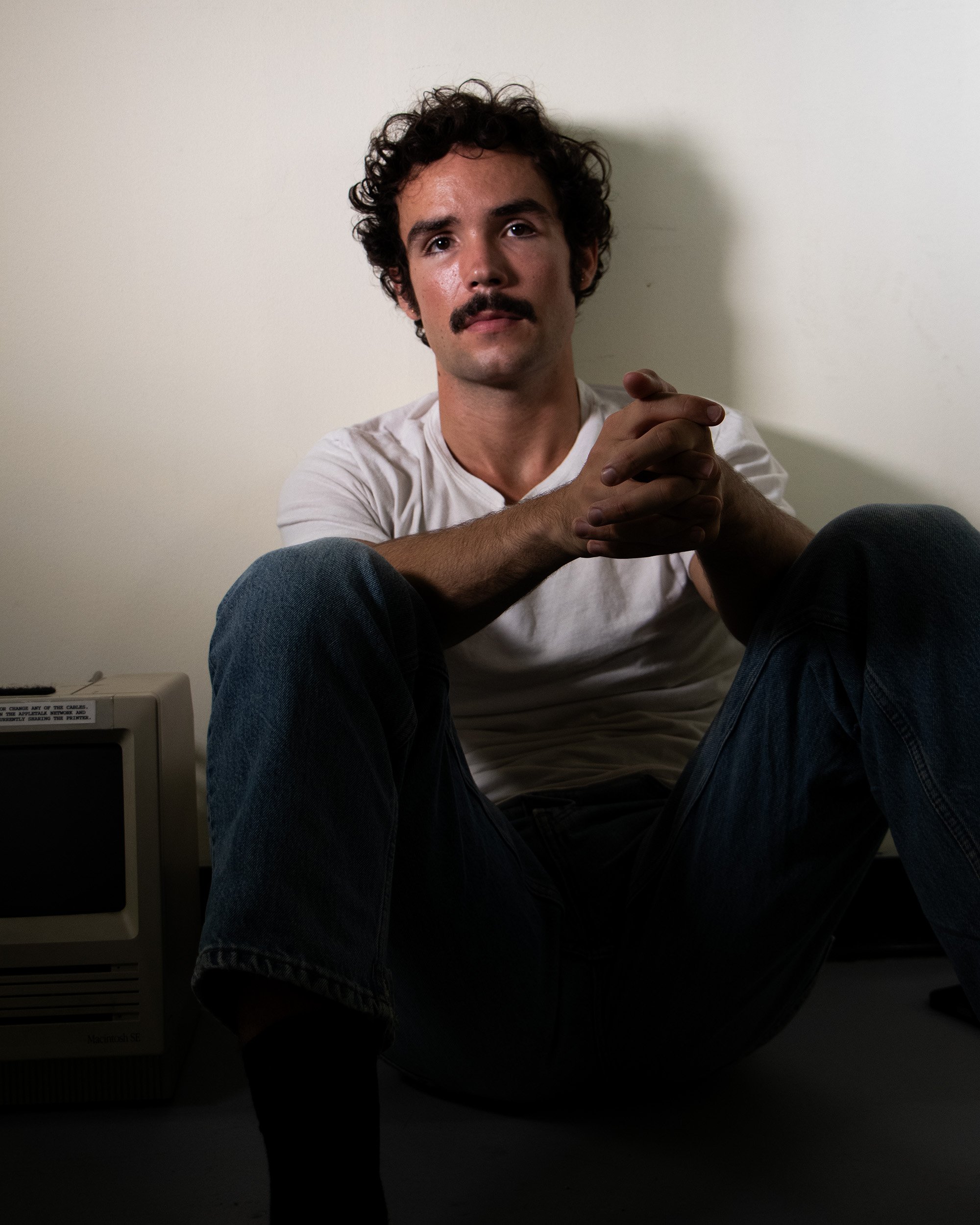 A young man with curly dark hair, a mustache, and light skin, sitting on the floor against a plain white wall, wearing a white t-shirt and jeans, with his hands clasped in front of him.