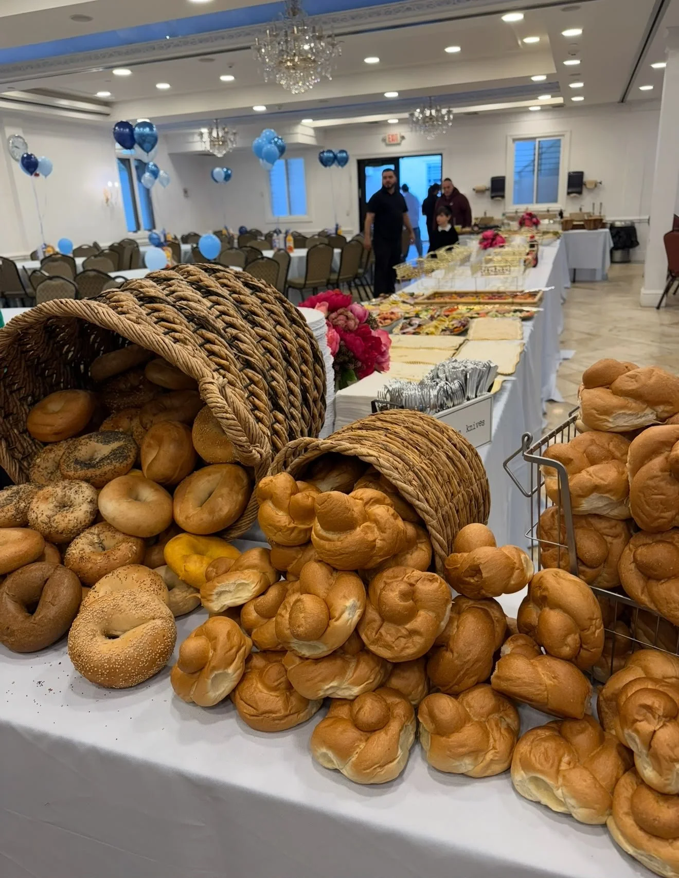 Display of assorted bread and bagels on a table at an event with decorations and a buffet setup in the background.