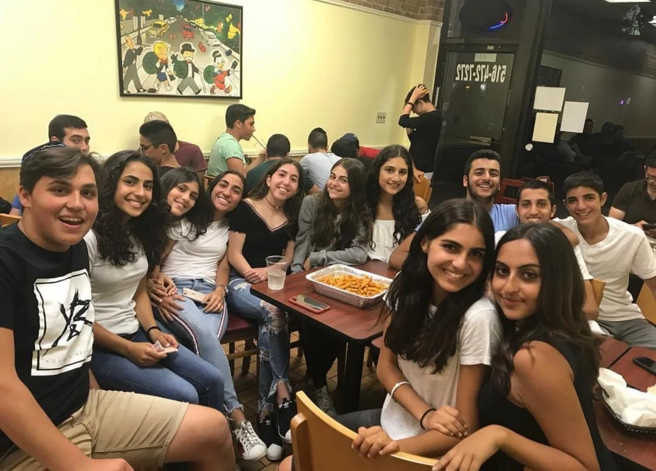 A group of young people sitting around a table in a restaurant, smiling at the camera, with a tray of food in front of them.