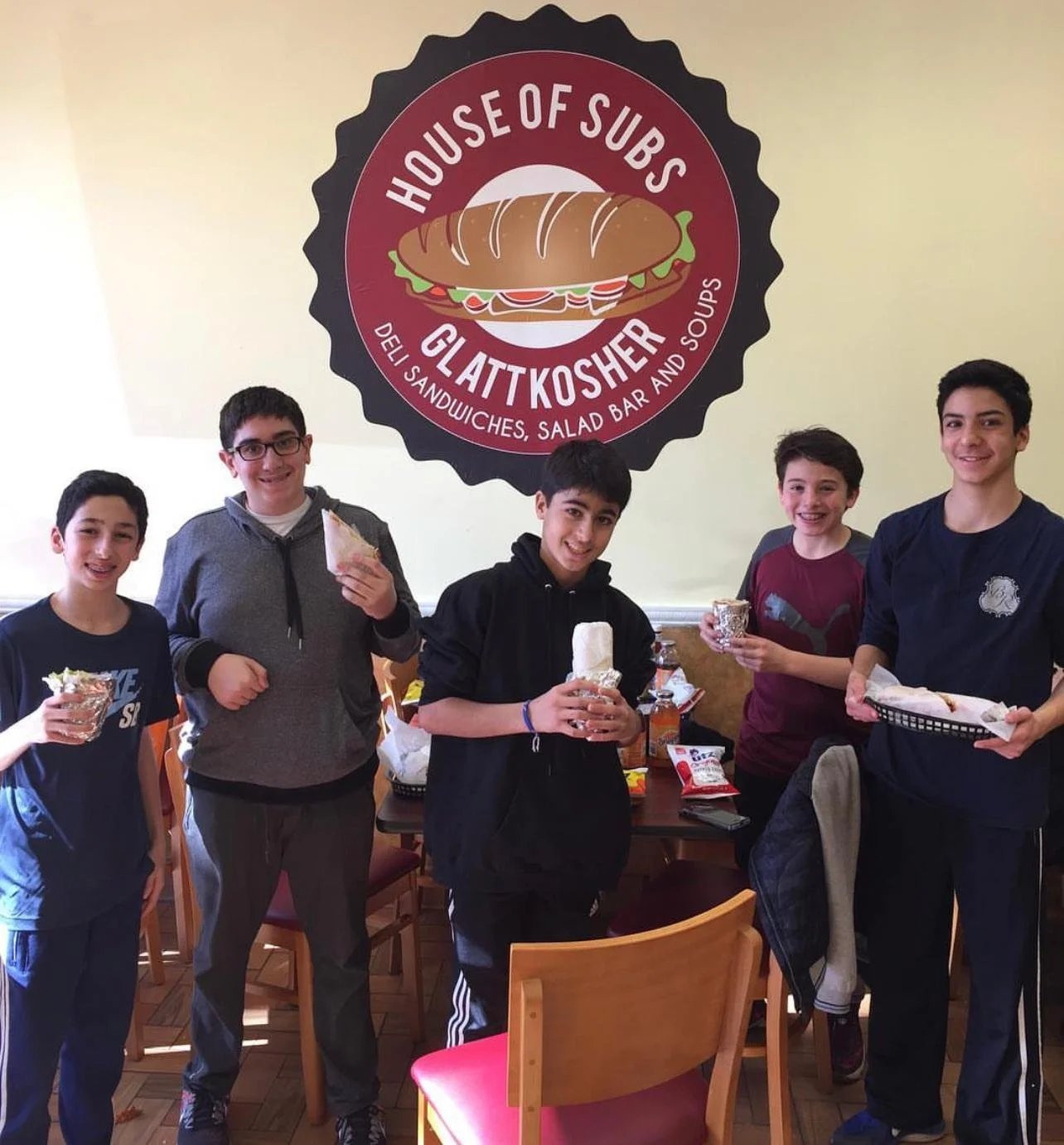Group of five boys standing in a restaurant holding food and drinks, with a large sign behind them that says 'House of Subs' and 'Glatt Kosher'.