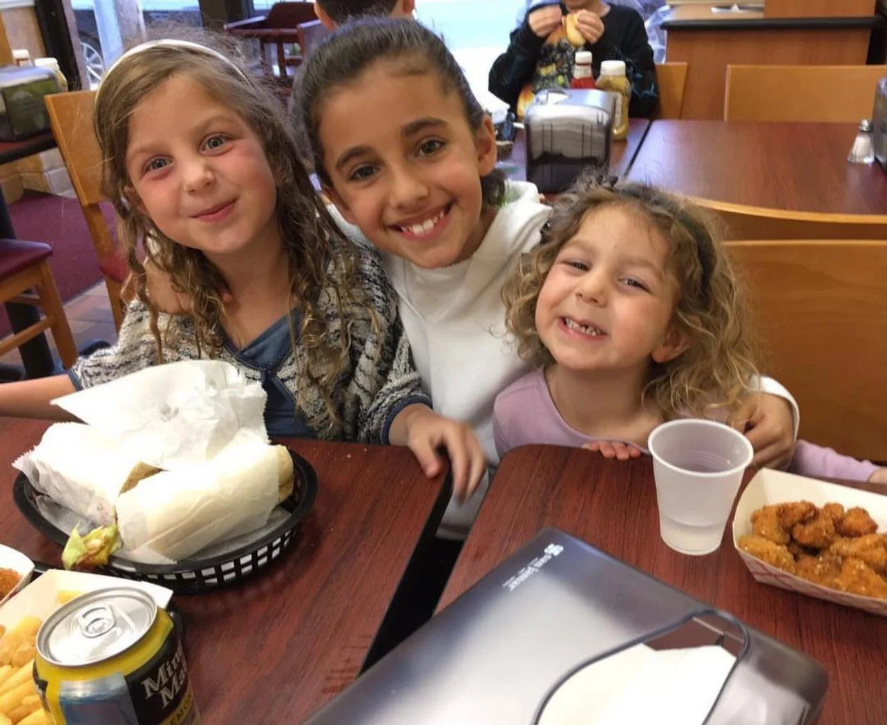 Three young girls smiling happily at a restaurant table with food and drinks, including sandwiches and fried chicken.