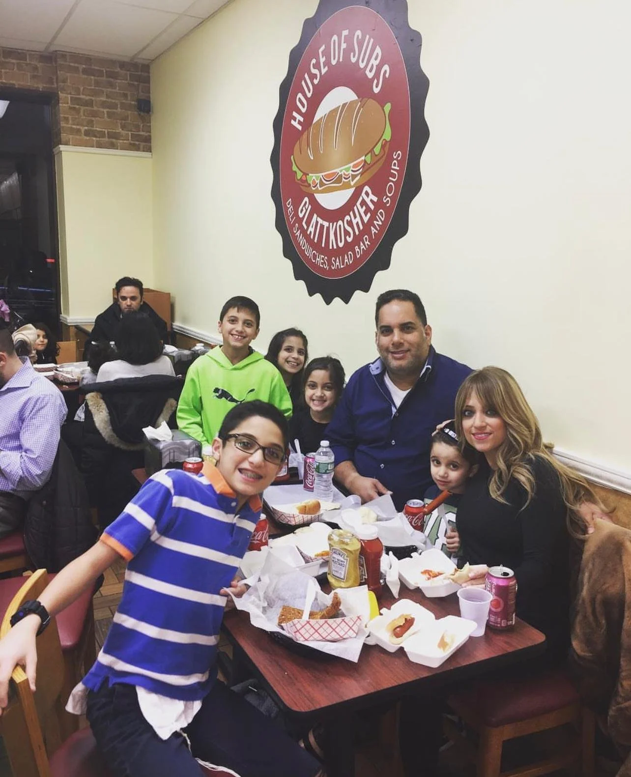 Family celebrating at a restaurant with tables of food, drinks, and condiments, and a large logo on the wall reading 'House of Subs Glaütkosher' with a sandwich illustration.