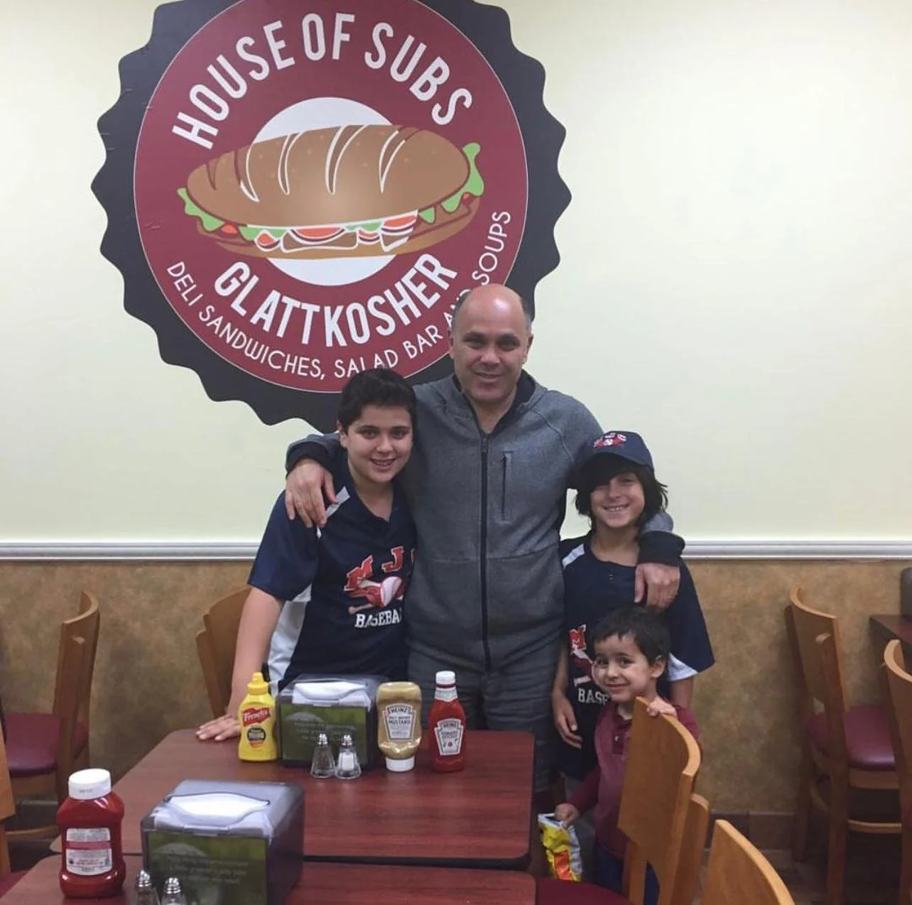 A group of five people, including a man and four children, standing at a restaurant table with condiments, in front of a logo for 'House of Subs Gluten Kosher' with an illustration of a sandwich. The man has his arms around two children, and two youn