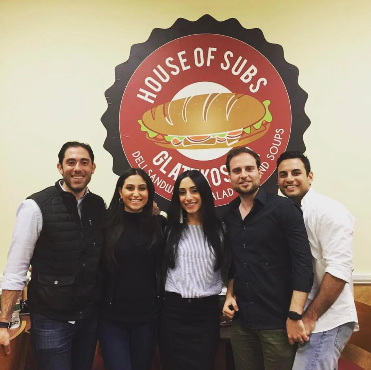 Five people standing together inside a restaurant, smiling for the photo, in front of a large circular sign that reads 'House of Subs Gluten-Free Sandwiches and Soups.'
