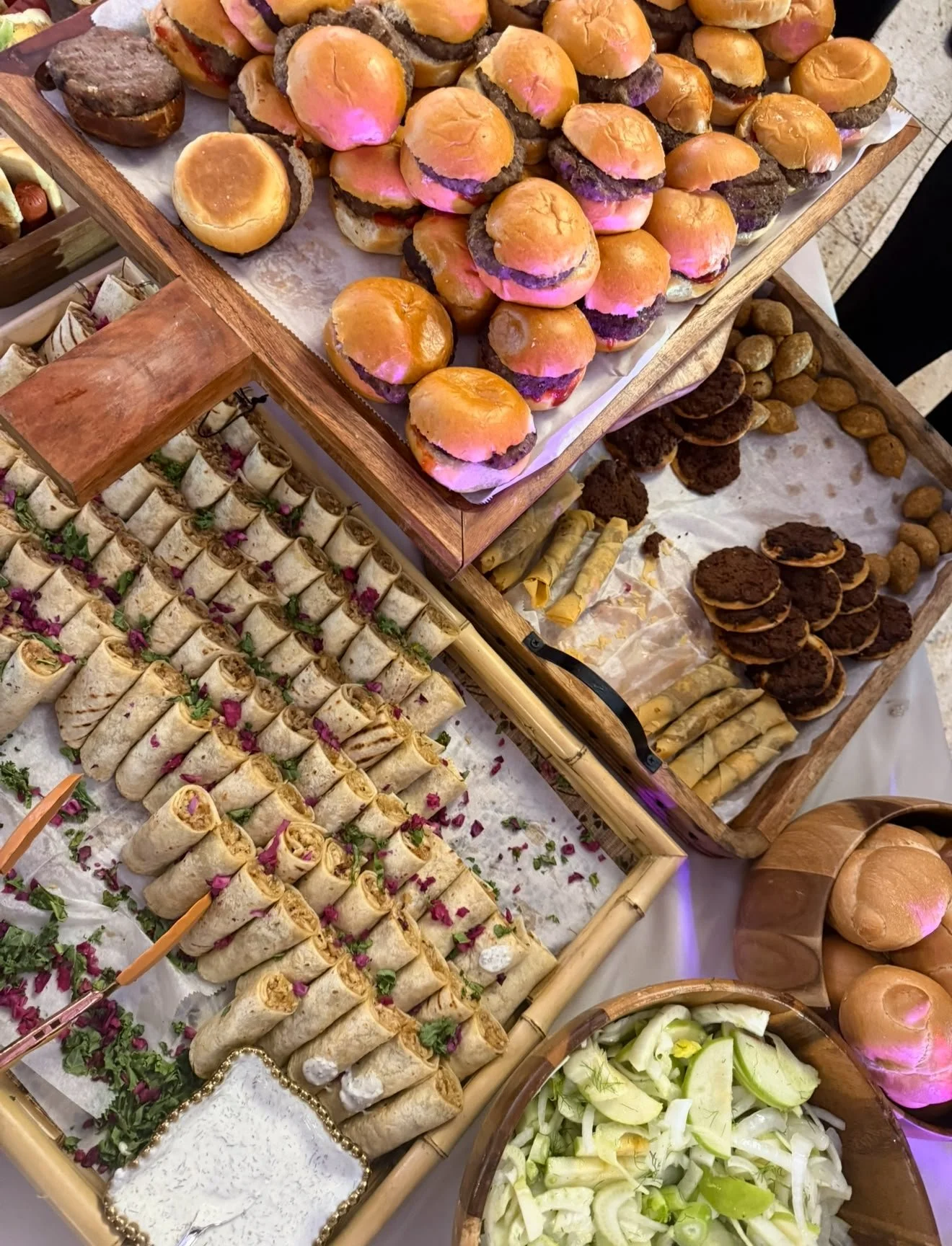 Assorted appetizers including mini burger sliders, rolled wraps with pink flower petals, and various cookies and chips on a buffet table.