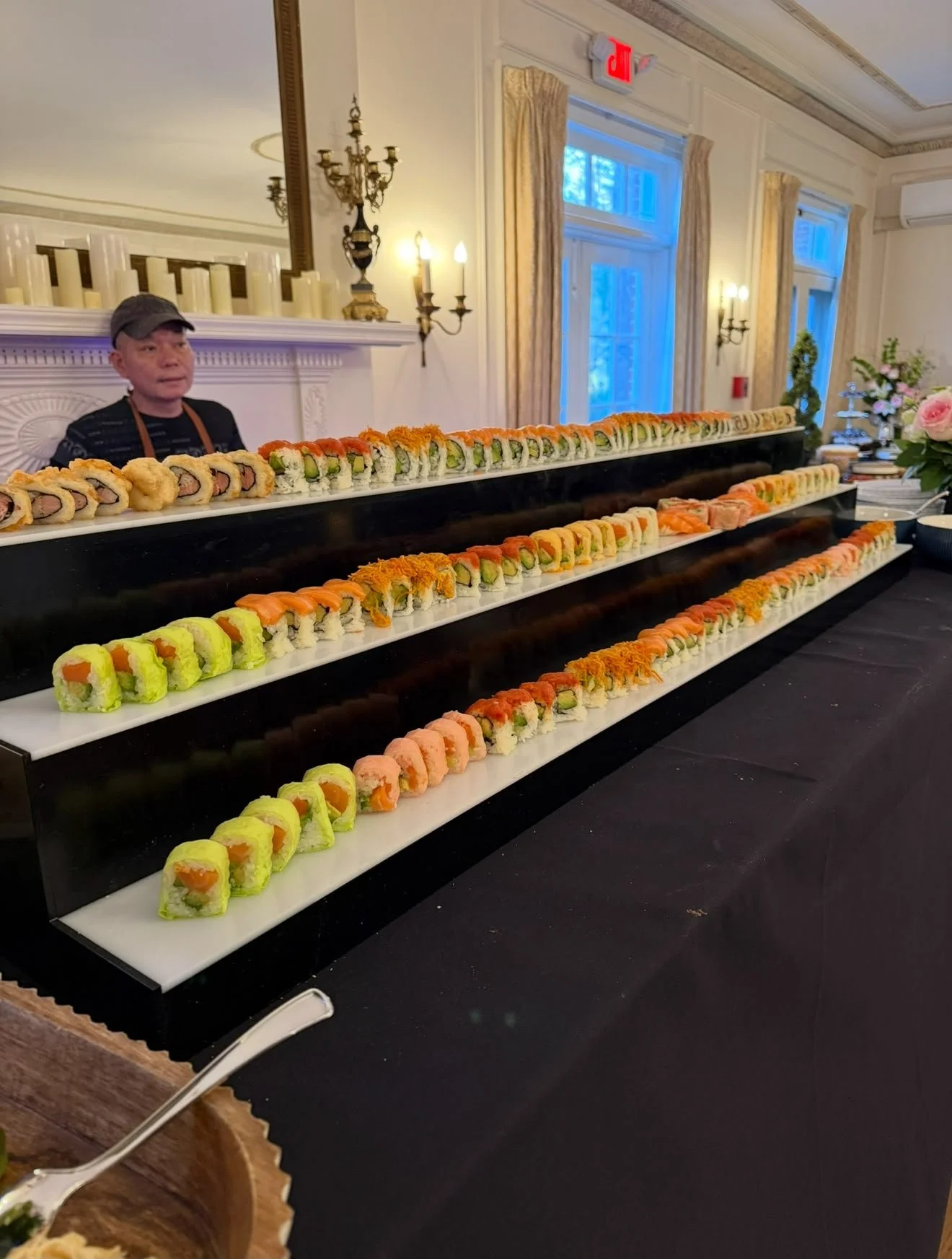 A man standing behind a long display of assorted sushi rolls arranged on white trays in a decorated room with large windows, beige curtains, and chandeliers.