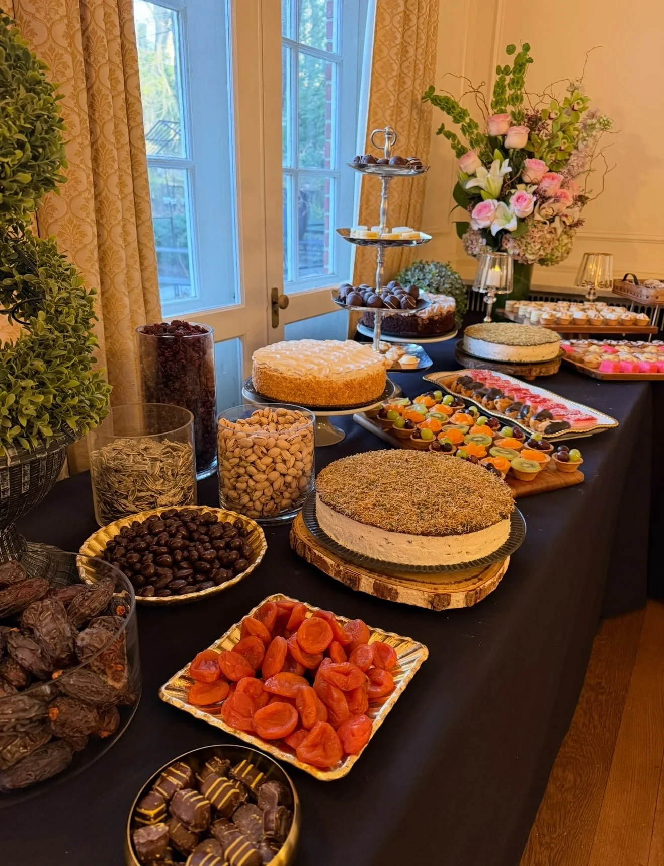 A table with various desserts, including cakes, chocolates, candies, and cookies, decorated with flowers near a window.